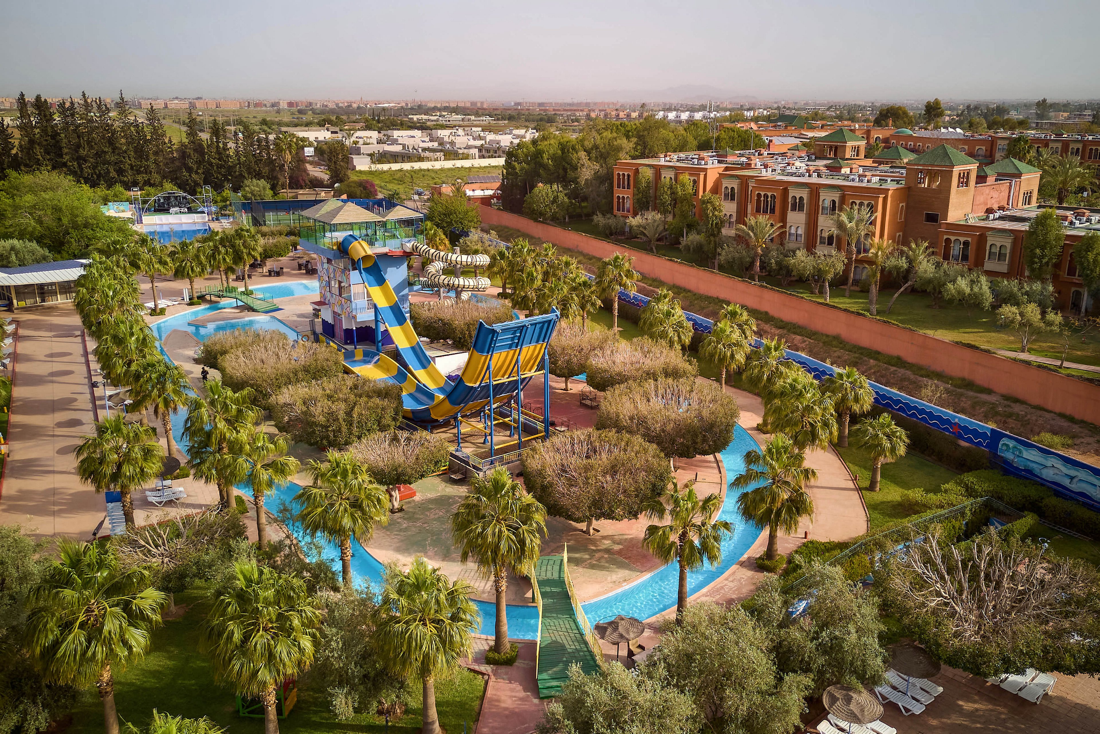 A large outdoor water park is captured from above, featuring a prominent blue and yellow slide structure. The slide curves into a winding lazy river surrounded by palm trees and landscaped greenery. Nearby, resort-style buildings with terracotta tones border the park, separated by a long red wall. The scene suggests a sunny, warm climate and a relaxed vacation atmosphere.
