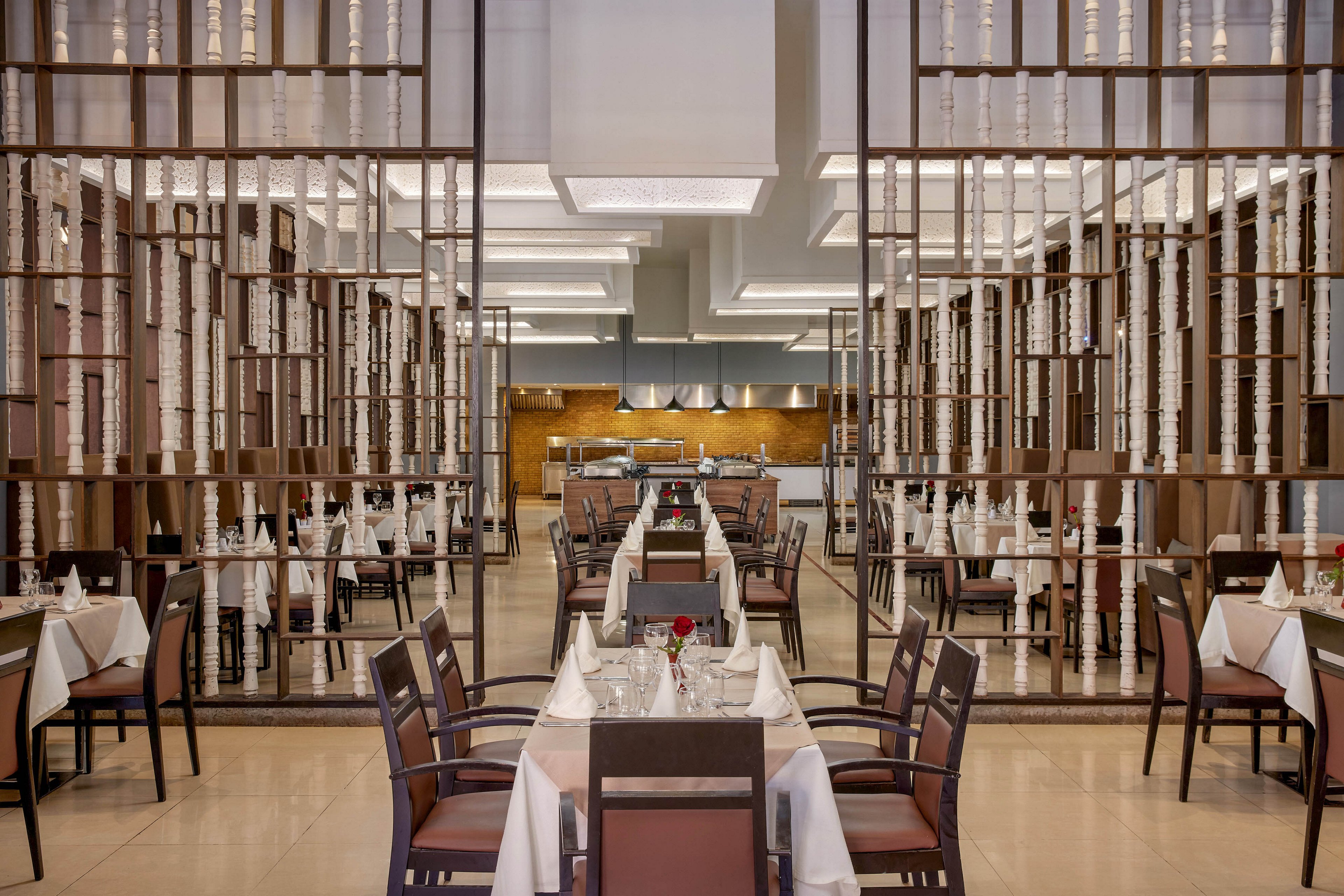 A contemporary restaurant dining room features neatly arranged tables with white tablecloths and brown chairs. Each table is set with glassware, cutlery, folded napkins, and a single red flower centerpiece. Decorative wooden partitions with vertical elements frame the space, leading the eye toward an open kitchen area at the back. Soft, even lighting and clean lines create a refined, welcoming atmosphere suitable for fine dining.