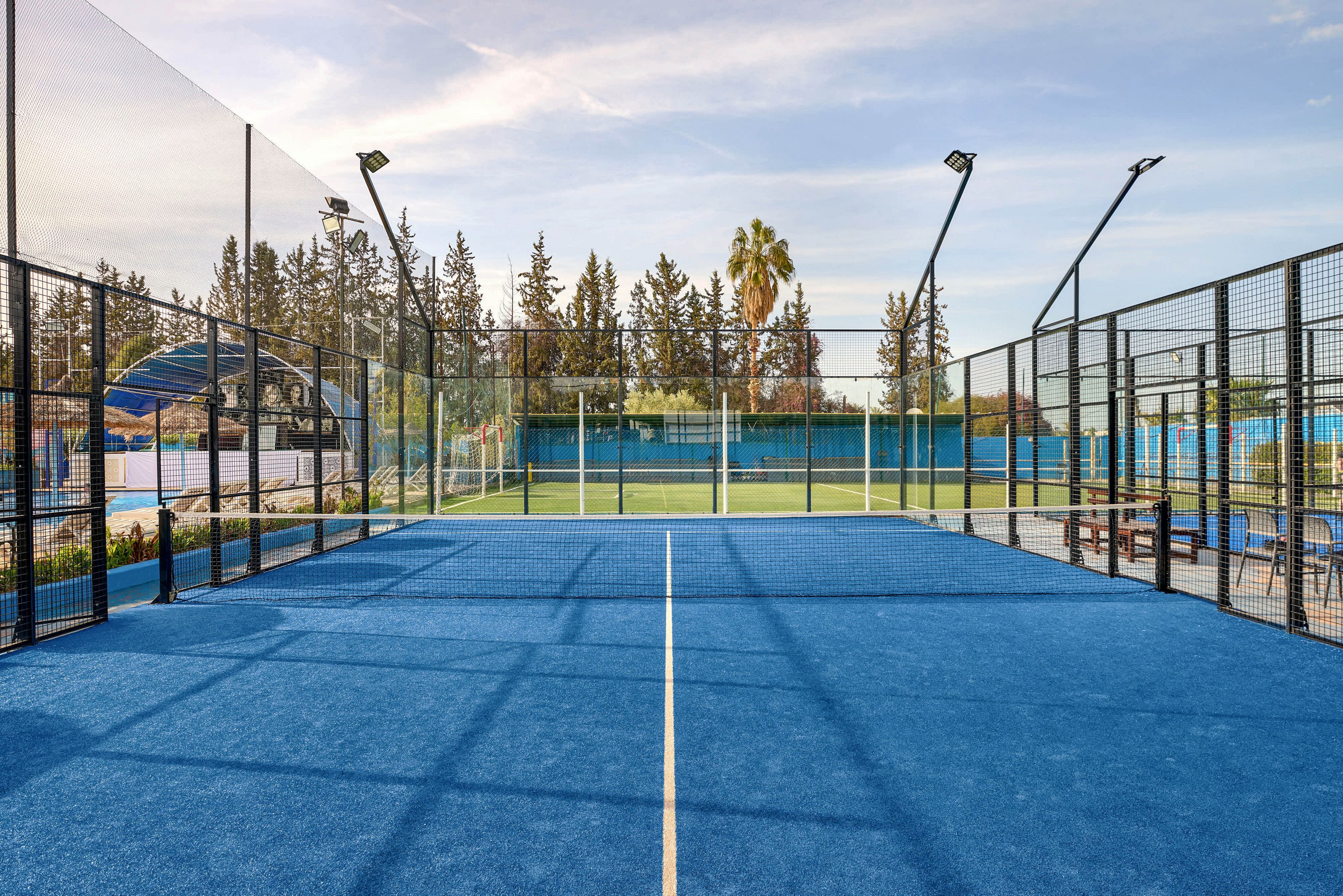 A bright blue padel tennis court is shown from one end, framed by tall glass and metal fencing. The court is outdoors, surrounded by trees and additional sports facilities in the background. Overhead lights and netting indicate a professional recreational setting under a clear sky. Benches and side structures suggest a club or sports complex environment ready for players.