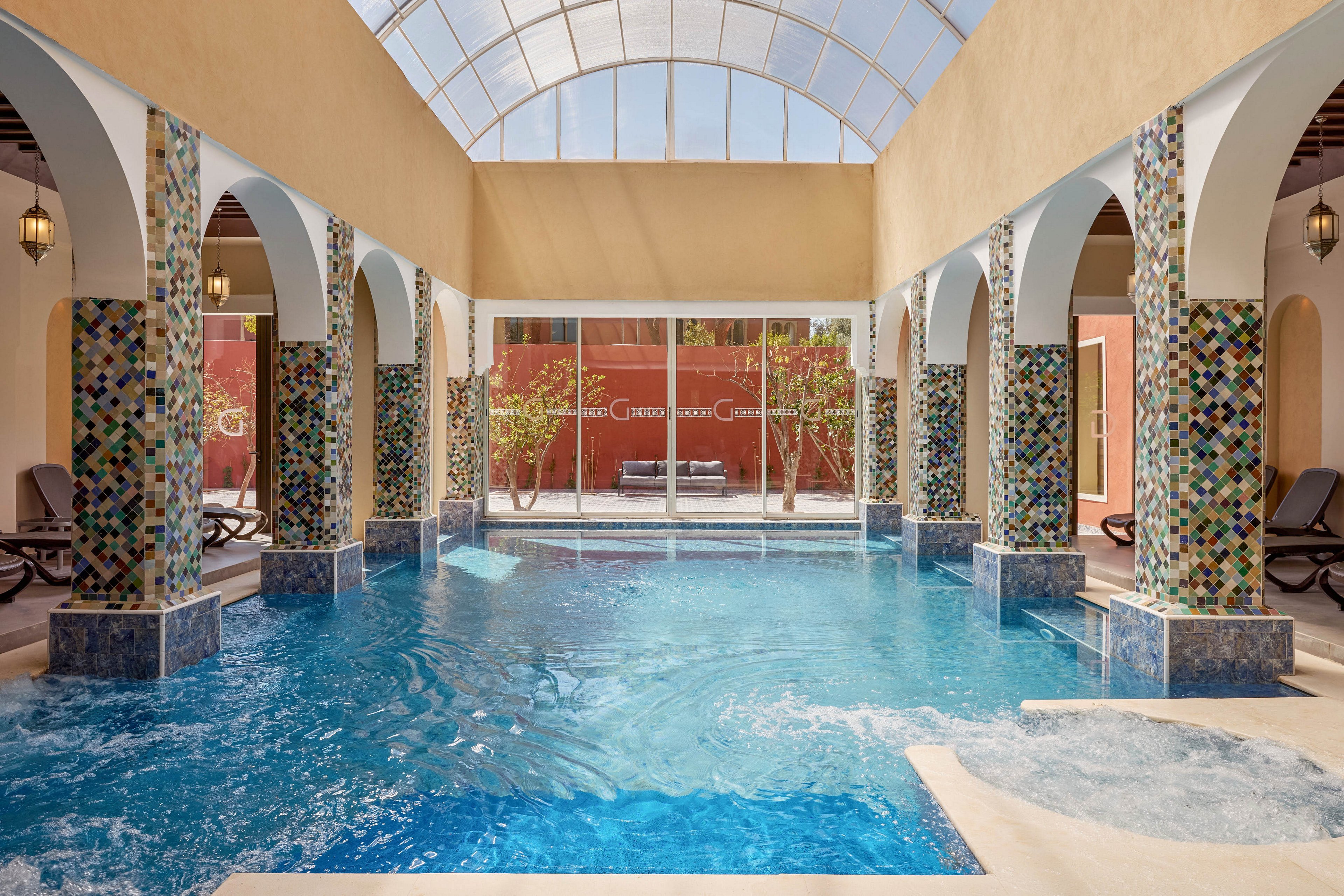 An inviting indoor pool is surrounded by arched columns decorated with colorful mosaic tiles. Lounge chairs line the sides of the pool area, suggesting a relaxing spa or resort setting. Natural light streams in through a large glass wall and a curved skylight above, illuminating the blue water. Outside the glass, a courtyard with trees and seating is visible, enhancing the tranquil atmosphere.