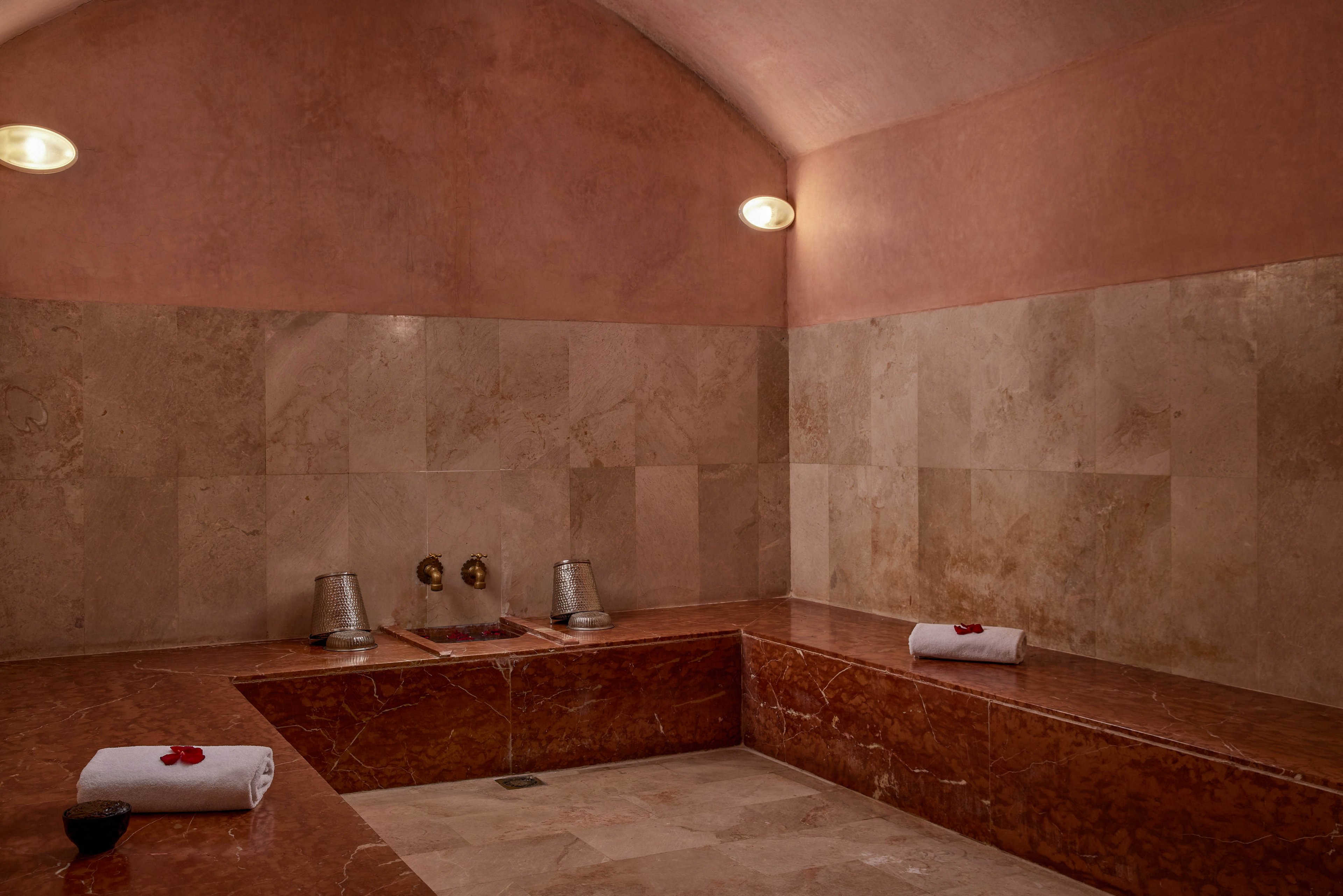 A spacious steam room features warm-toned stone benches and walls under a softly arched ceiling. Two rolled white towels with red flower petals rest on the polished stone surfaces, suggesting a prepared spa experience. Metal buckets and wall-mounted faucets sit at the center, indicating traditional bathing or rinsing rituals. Soft wall lights create a calm, intimate atmosphere ideal for relaxation and wellness.
