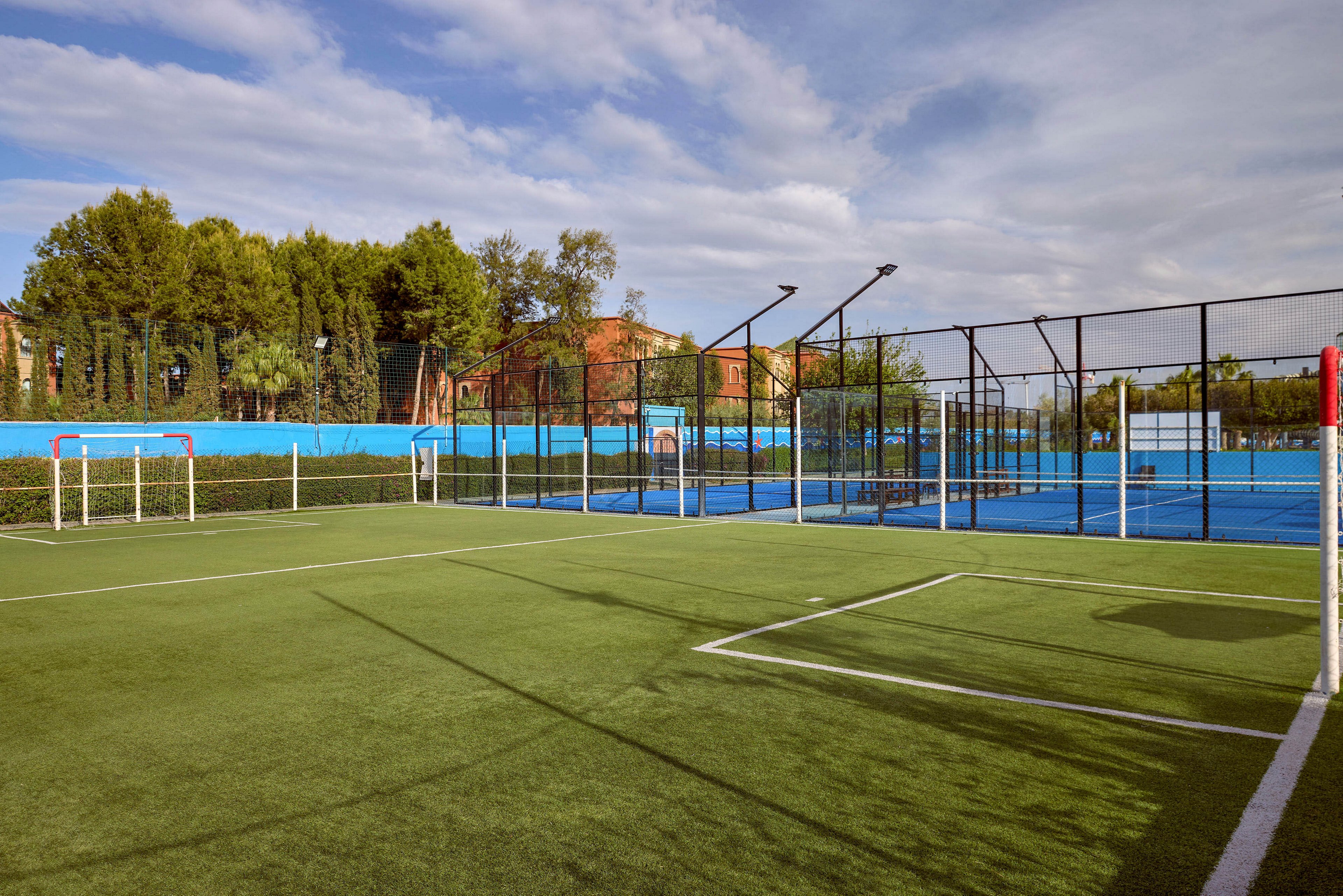 An outdoor sports complex features a small soccer field in the foreground with mini goals on artificial turf. Behind it, fenced blue courts are visible, likely used for tennis or similar racket sports. Tall netting and metal fencing surround the courts, with trees and buildings in the background. The scene is captured under a partly cloudy sky, suggesting a calm day at the recreational facility.
