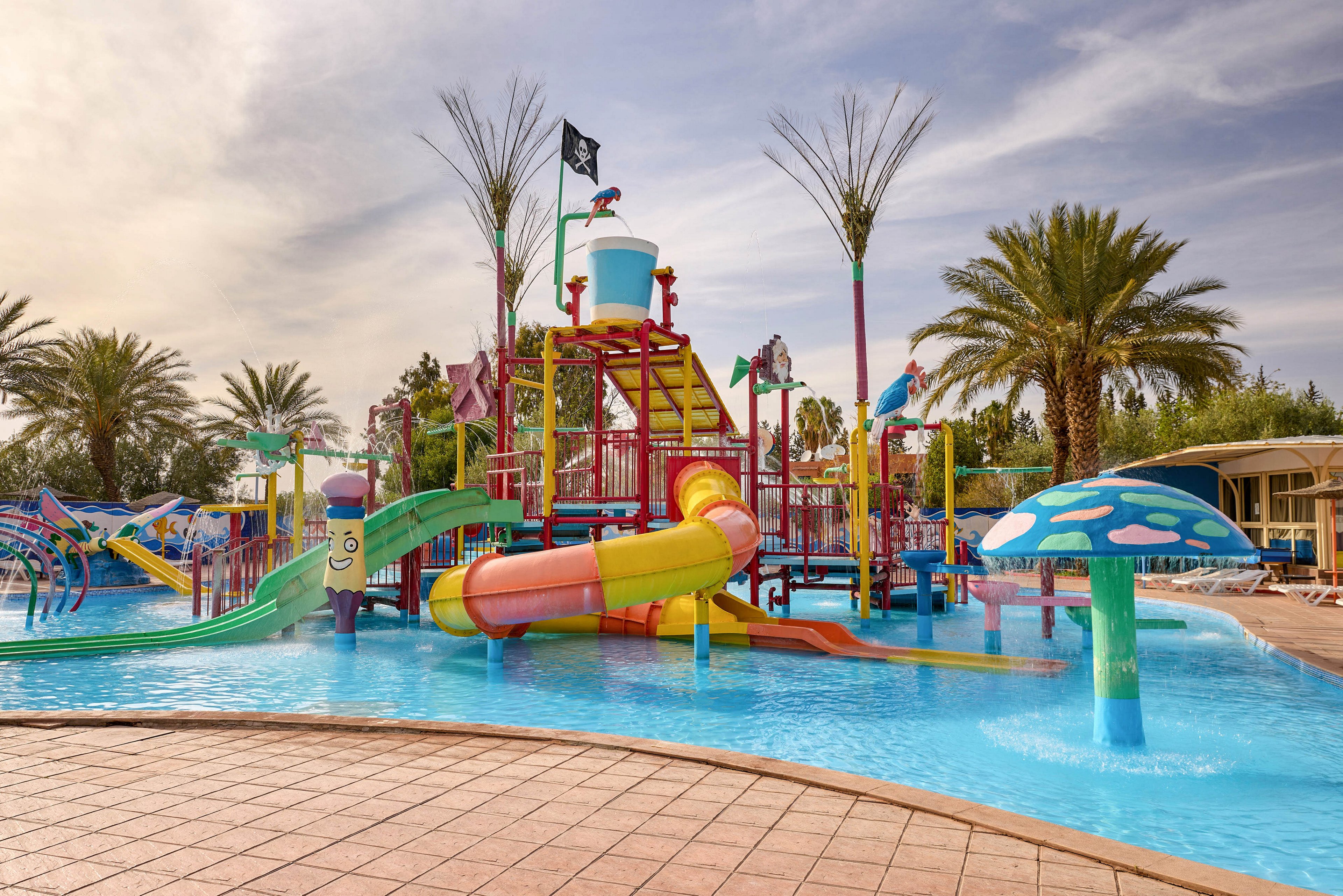 A vibrant water playground features multiple colorful slides and splash structures in a shallow pool. Palm trees and landscaped greenery surround the area, creating a tropical resort atmosphere. A large tipping bucket, water sprayers, and mushroom-shaped fountains add playful visual interest. The scene appears calm and empty, suggesting early morning or off-peak hours at the water park.