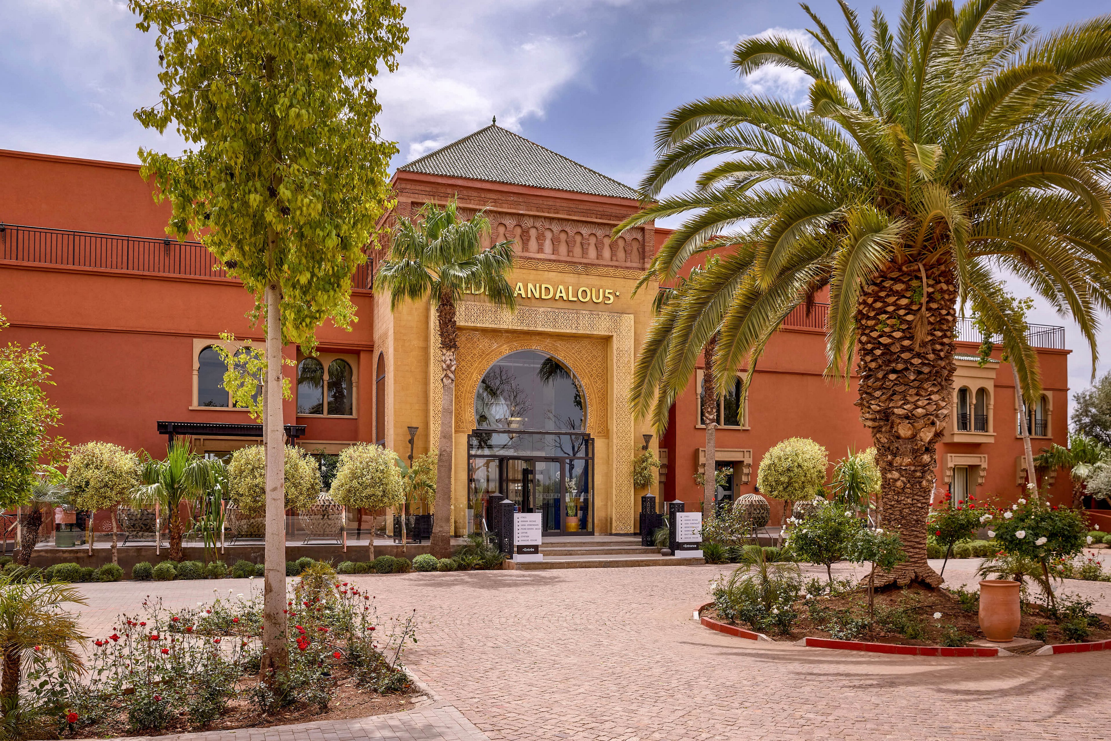 A large building with the name ANDALOUS above a grand arched entrance is shown in a sunny courtyard. Tall palm trees and manicured shrubs frame the pathway leading to the glass doors. The architecture features warm terracotta walls and decorative golden tones around the main archway. The scene suggests a welcoming hotel or resort environment in a warm climate.