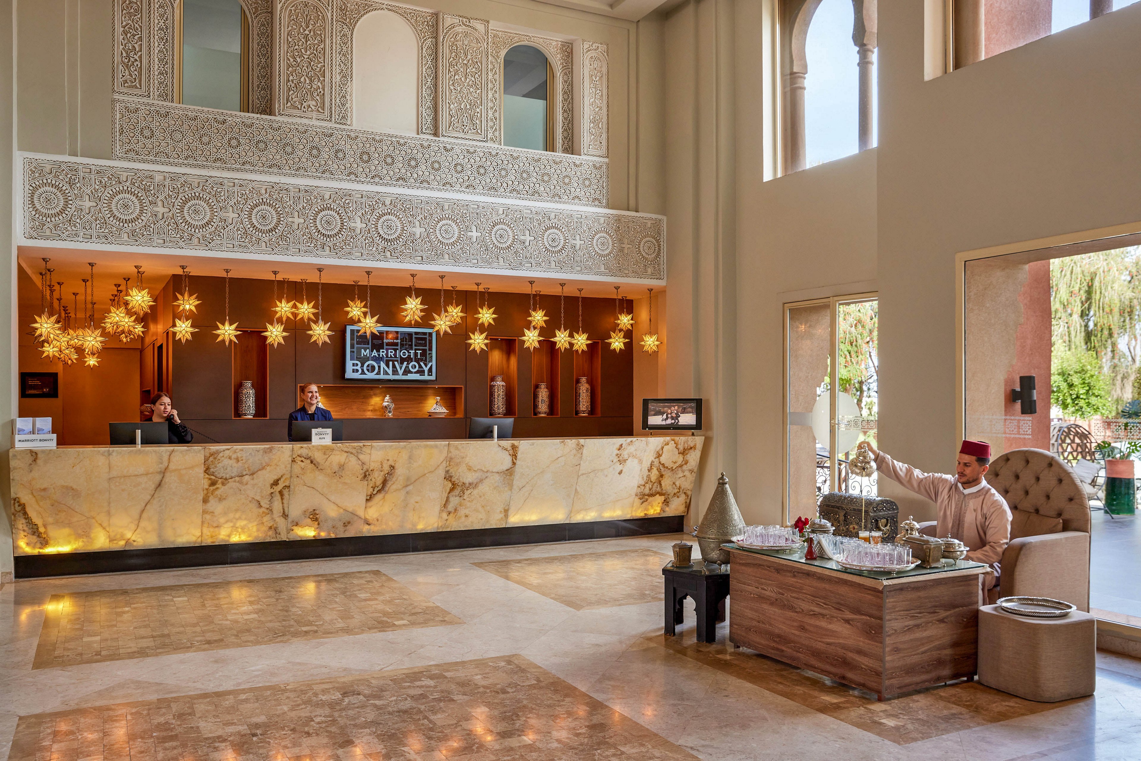 A spacious hotel lobby features a marble-front reception desk with staff attending guests. Ornate star-shaped pendant lights and intricate wall patterns create a luxurious, contemporary Middle Eastern-inspired interior. On the right, a man in traditional attire serves tea from a silver set at a low wooden table, adding a welcoming cultural touch. Large windows reveal greenery outside, enhancing the bright and inviting atmosphere.