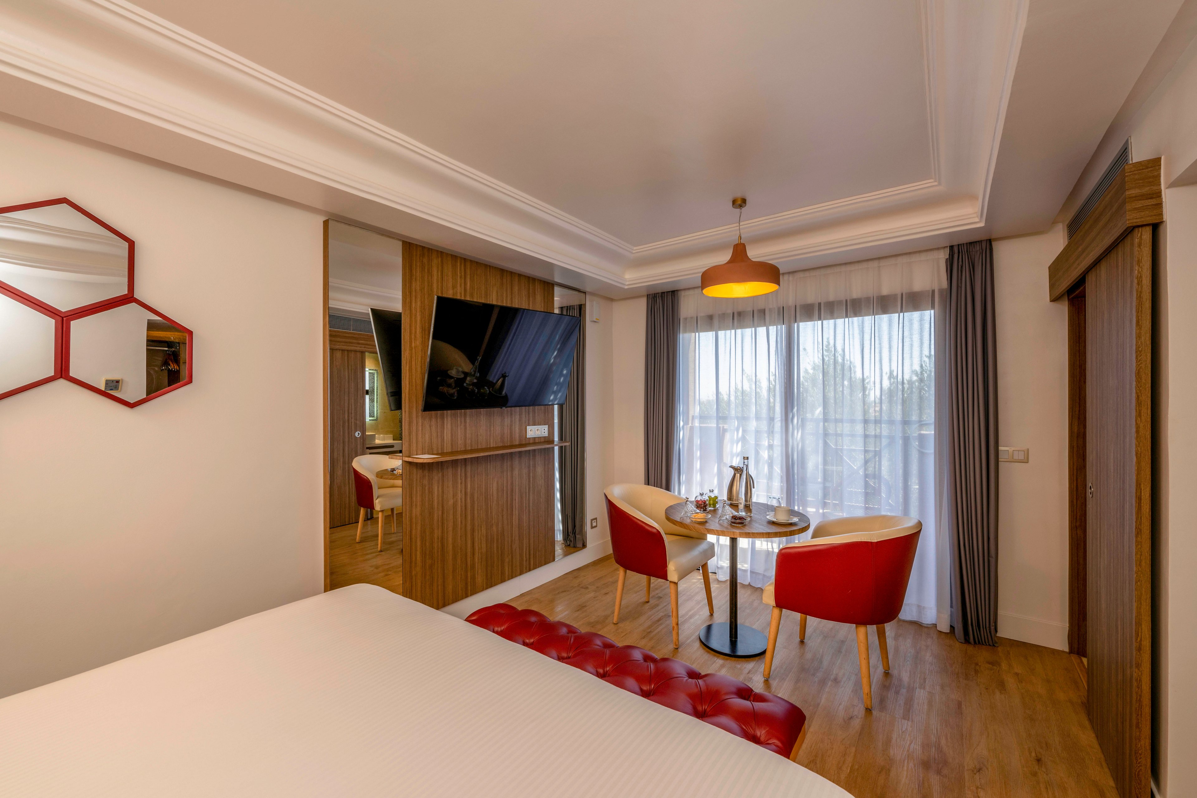A contemporary hotel room features a neatly made bed facing a wall-mounted flat-screen television. Near the large window with sheer curtains, a small round table is set with a metallic ice bucket and glasses, flanked by two red and beige chairs. Warm wooden flooring and paneling complement the white ceiling and walls, while a geometric red-framed mirror adds a decorative touch. Soft ambient lighting from a ceiling pendant creates an inviting and comfortable atmosphere.