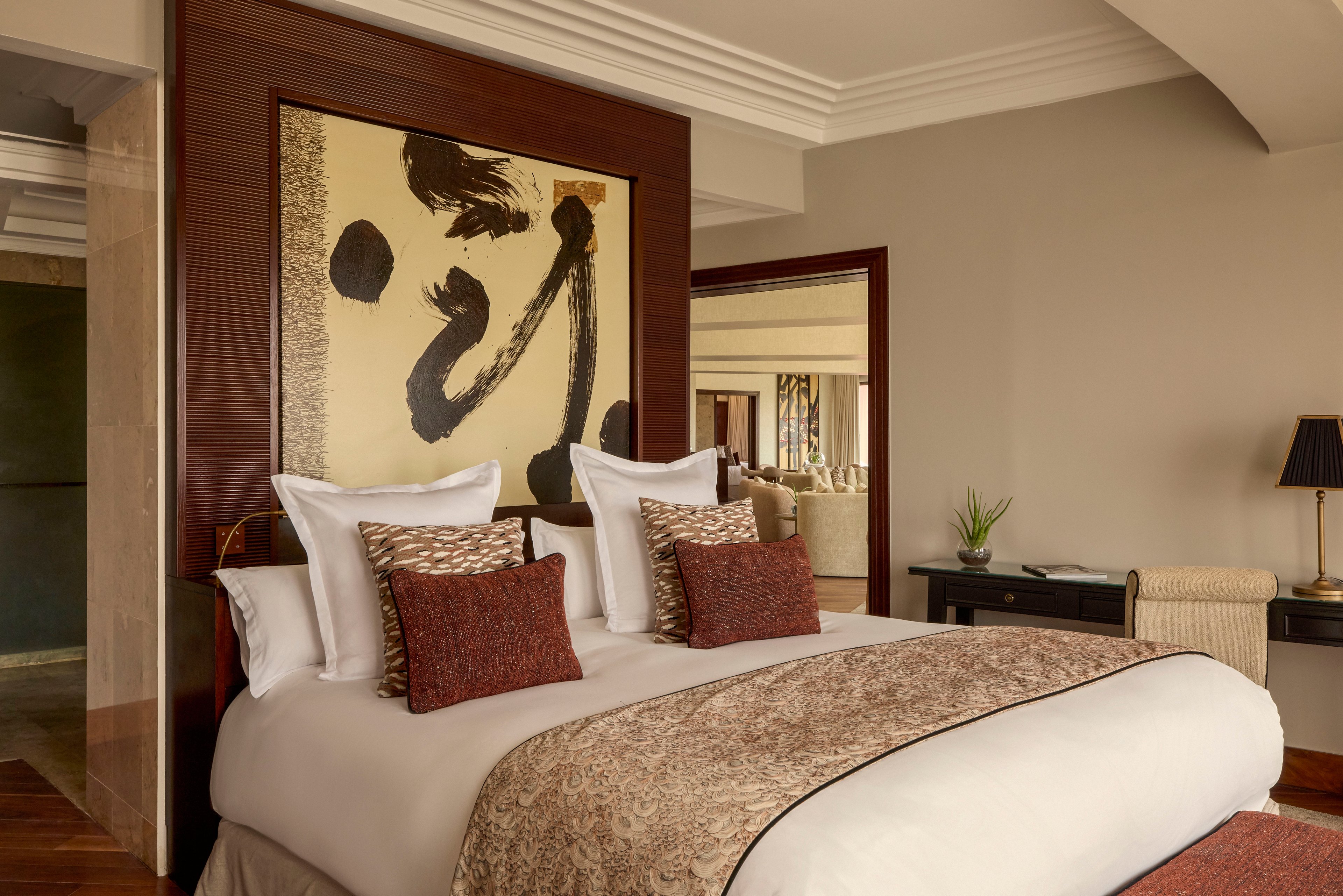 A neatly made double bed with plush pillows and a patterned throw blanket stands as the focal point of a stylish hotel bedroom. Warm beige walls and dark wood accents create a sophisticated, inviting atmosphere. Abstract artwork above the headboard adds a contemporary touch, while a side table with a lamp and a seating area are visible in the background. The overall scene suggests comfort, relaxation, and upscale interior design.