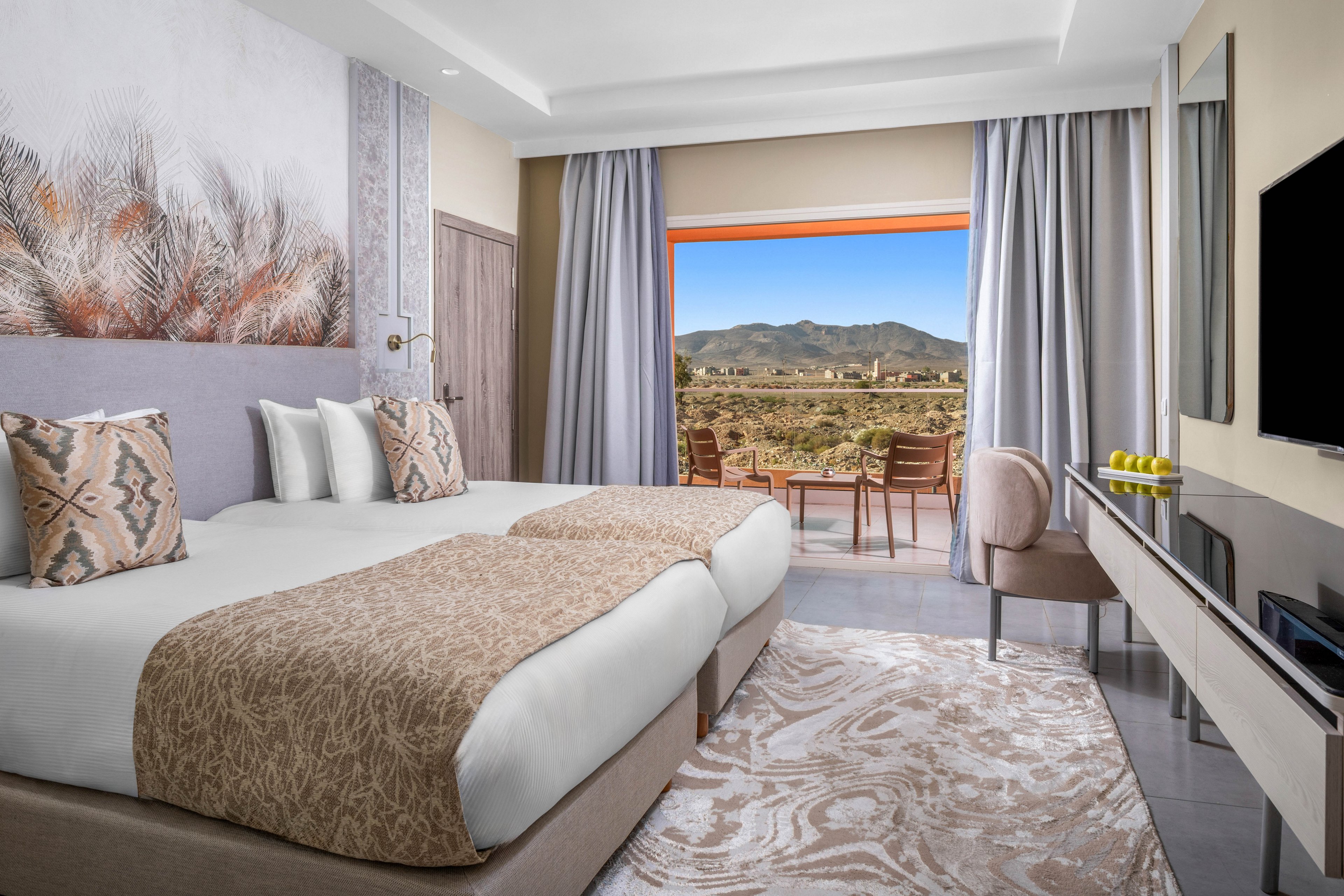 A contemporary hotel room features two twin beds with patterned bedding and decorative pillows. The space is styled in neutral tones with a nature-inspired wall mural and a large window opening to a balcony overlooking mountains. The room includes a desk, television, and a bowl of green apples, creating a welcoming and serene atmosphere.