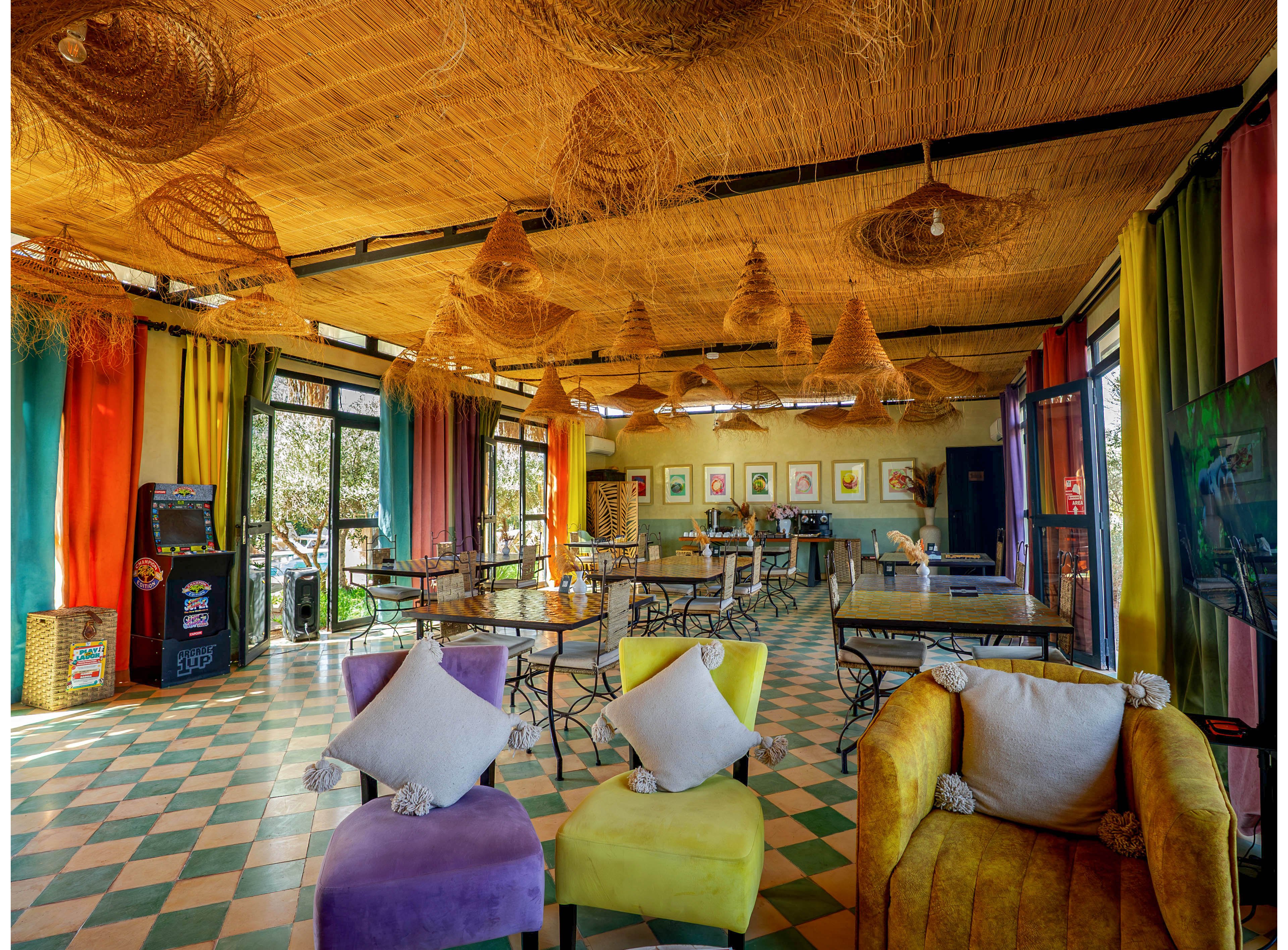 Vibrant cafe interior featuring colorful armchairs and wicker light fixtures hanging from a straw-covered ceiling. The space includes several metal-framed tables and chairs arranged on a green and cream checkered tile floor. Large windows with multicolored curtains allow natural light to fill the room and reveal greenery outside. Framed artwork on the back wall and a vending machine in the corner add to the eclectic, relaxed atmosphere.