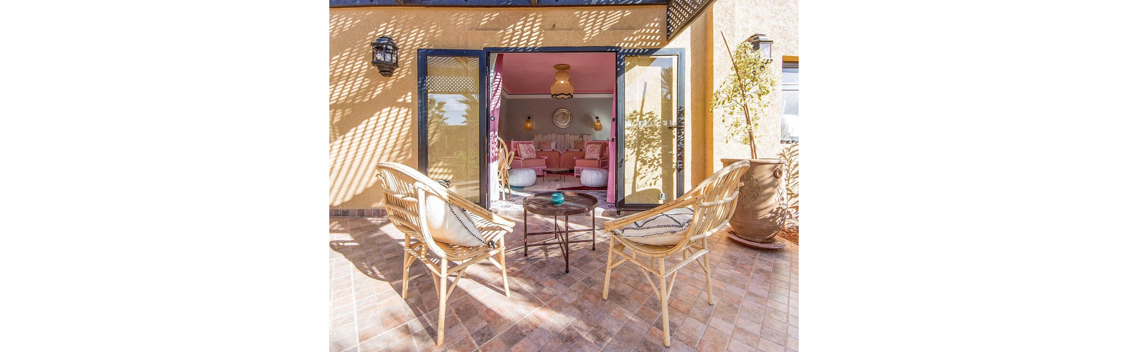 Outdoor wicker chairs and a small round table sit on a tiled patio in bright sunlight. Glass doors open into a cozy indoor lounge with pink walls and cushioned seating. Decorative pendant lights and wall art add warmth and style to the interior. The scene suggests a relaxing retreat blending indoor comfort with outdoor living.