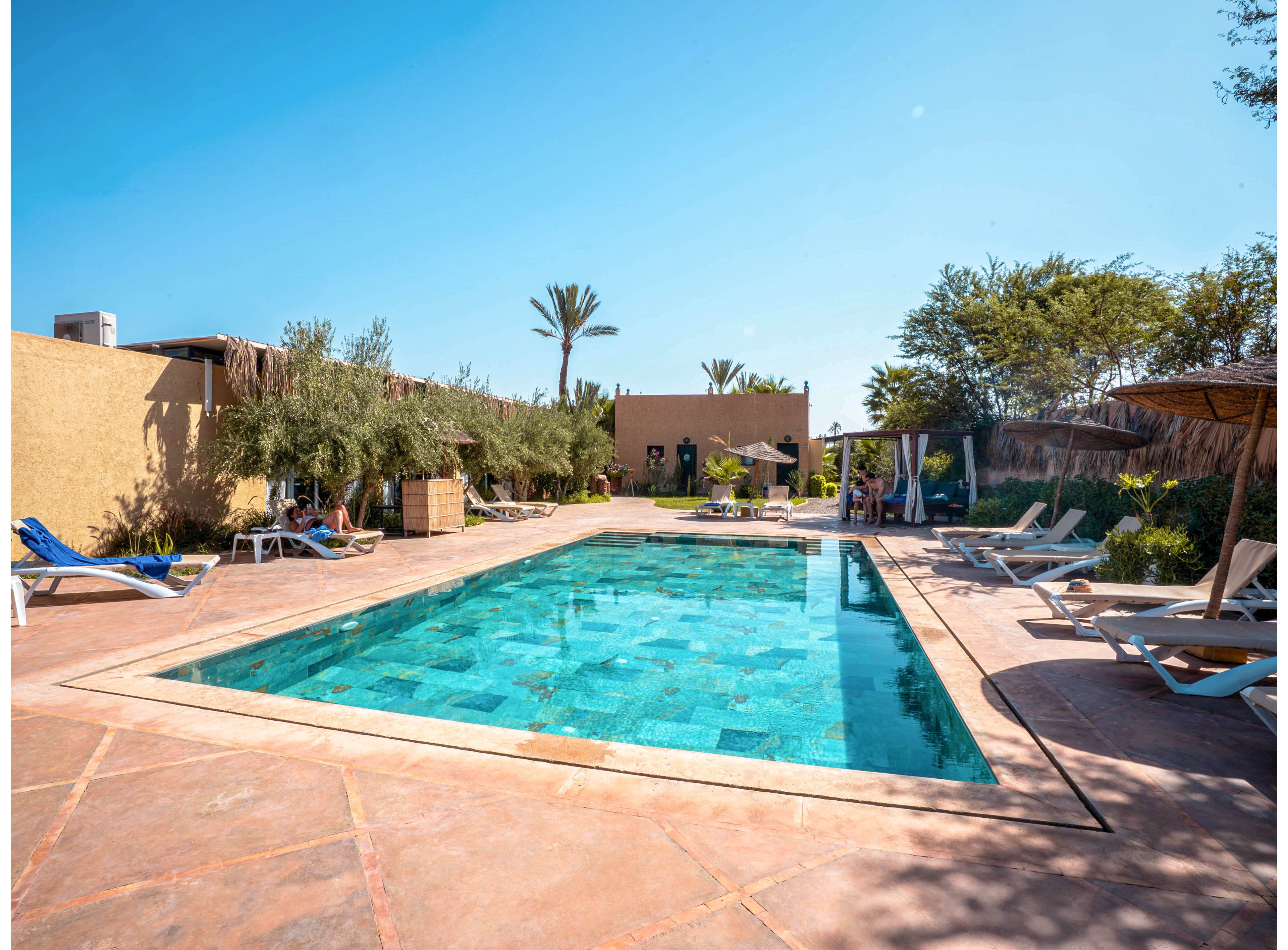 An outdoor swimming pool sits at the center of a sunny resort courtyard. Lounge chairs and umbrellas line the tiled pool deck, suggesting a relaxing vacation setting. Palm trees and lush greenery surround the area, with low buildings and clear blue sky in the background. The bright daylight emphasizes the turquoise water and warm stone surfaces, creating a tranquil holiday atmosphere.
