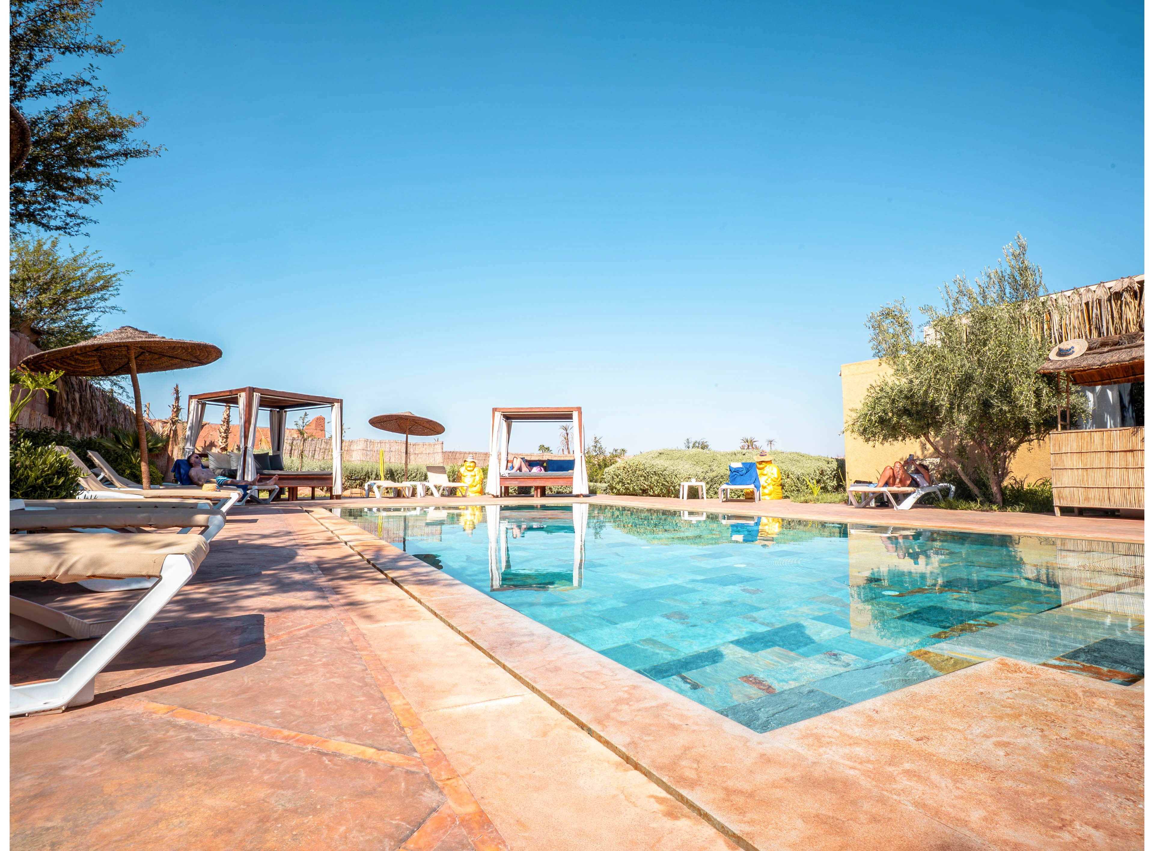 An outdoor swimming pool sits at the center of a sunny resort. Lounge chairs and shaded daybeds surround the pool, where a few people relax under umbrellas. The sky is clear and bright blue, and the water reflects the vivid color. Trees and low buildings frame the scene, creating a tranquil vacation atmosphere.