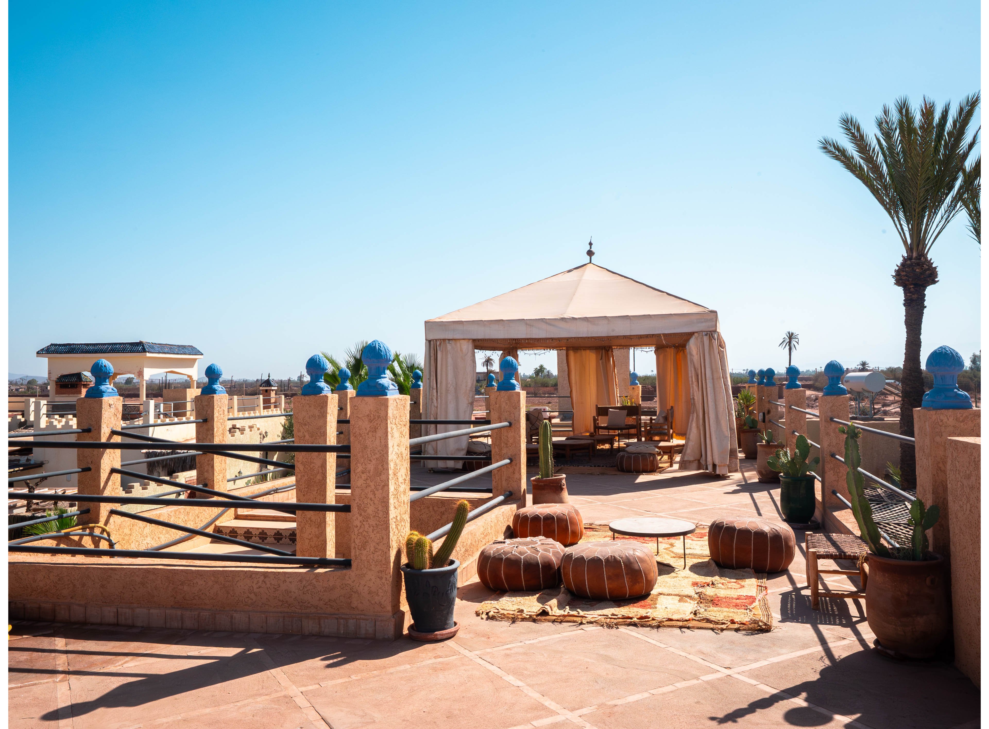 An inviting rooftop terrace features a central canvas tent with open sides and low seating. Round leather poufs and woven rugs create a cozy lounge area under the bright blue sky. Terracotta walls with blue-topped posts and potted cacti frame the space. A tall palm tree and distant rooftops suggest a warm, desert or Mediterranean city setting.