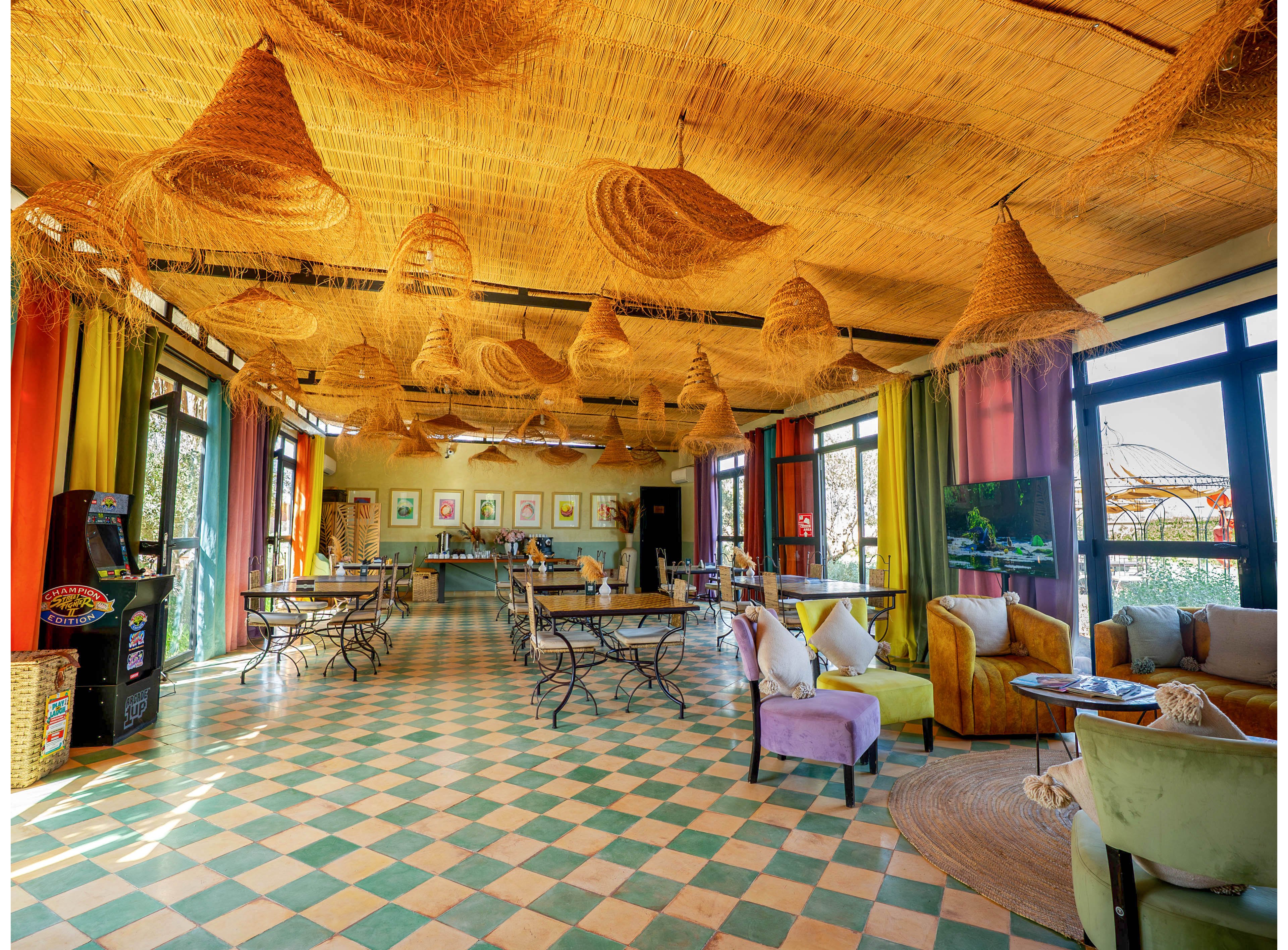 Bright, spacious cafe interior featuring wicker pendant lights hanging from a straw-covered ceiling. The room has checkered green and peach floor tiles and large windows letting in natural light. Colorful curtains and upholstered chairs in pastel tones create a cozy, eclectic atmosphere. Several small tables and chairs are arranged throughout the space, suggesting a relaxed dining or lounge area.