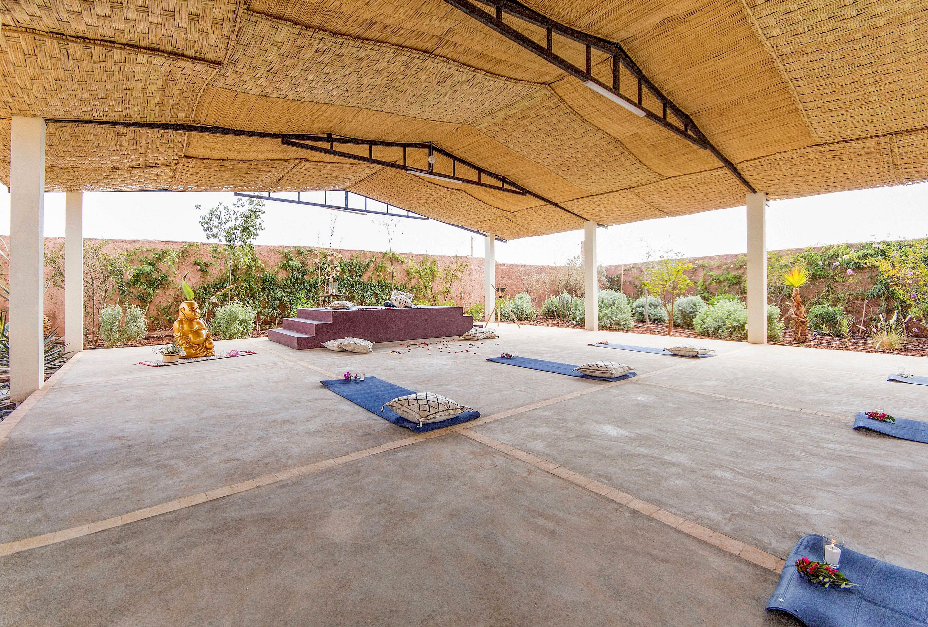 An open-air pavilion is arranged for a yoga or meditation session with mats laid out evenly on the floor. Each mat has a folded blanket and small decorative flowers placed on top. At the front of the space, a raised platform features a golden statue and stones arranged in a circular pattern. The structure has a woven, natural-looking roof and is surrounded by a garden with greenery and a tall wall, creating a calm, secluded atmosphere.