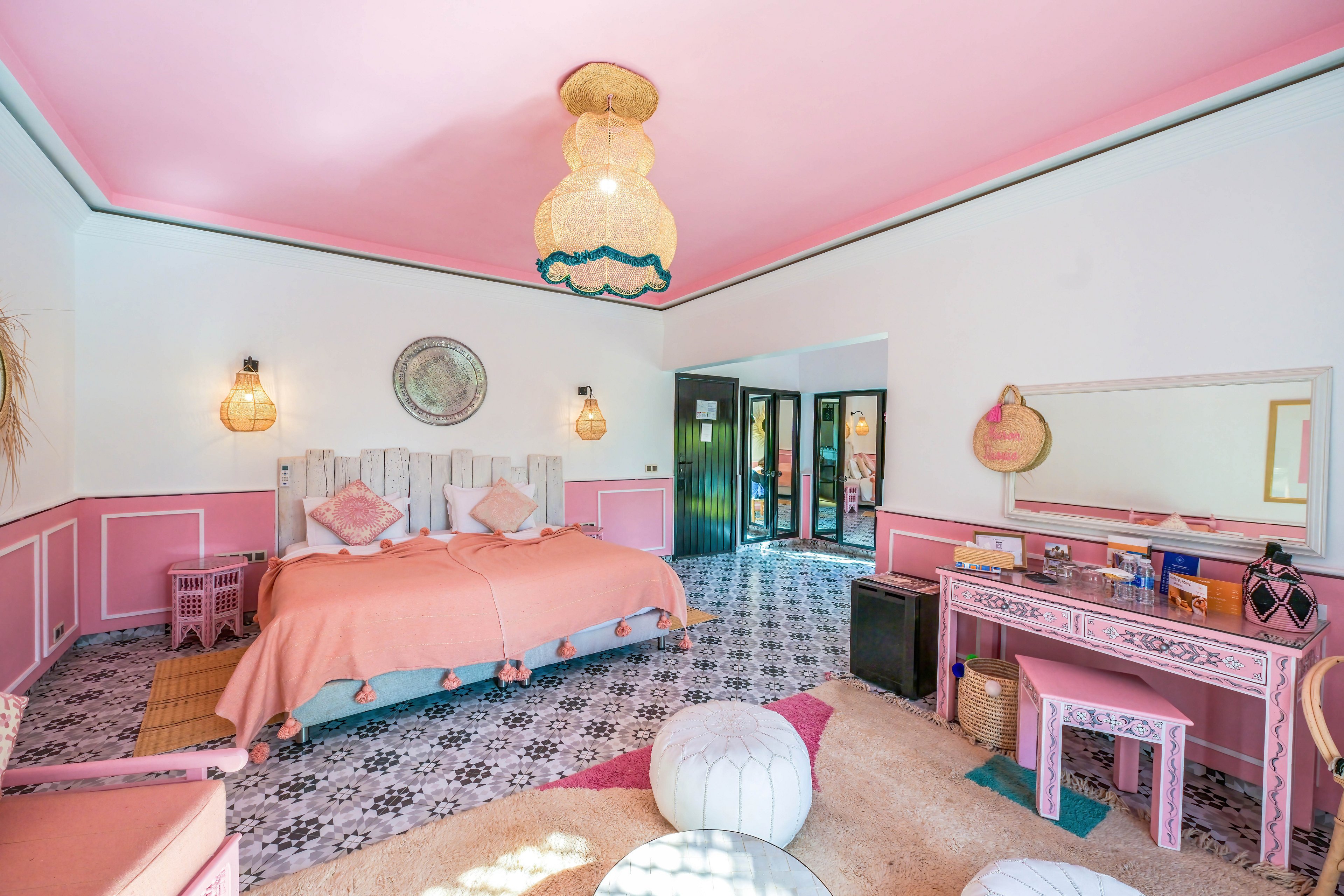 Brightly decorated bedroom with a large double bed features a playful pink and peach color scheme. The room includes patterned floor tiles, a vanity desk with mirror, and woven pendant lights. Natural light enters through glass doors leading outside, enhancing the airy, spacious feel. Decorative cushions, poufs, and textured rugs add a cozy, bohemian touch to the stylish hotel room.