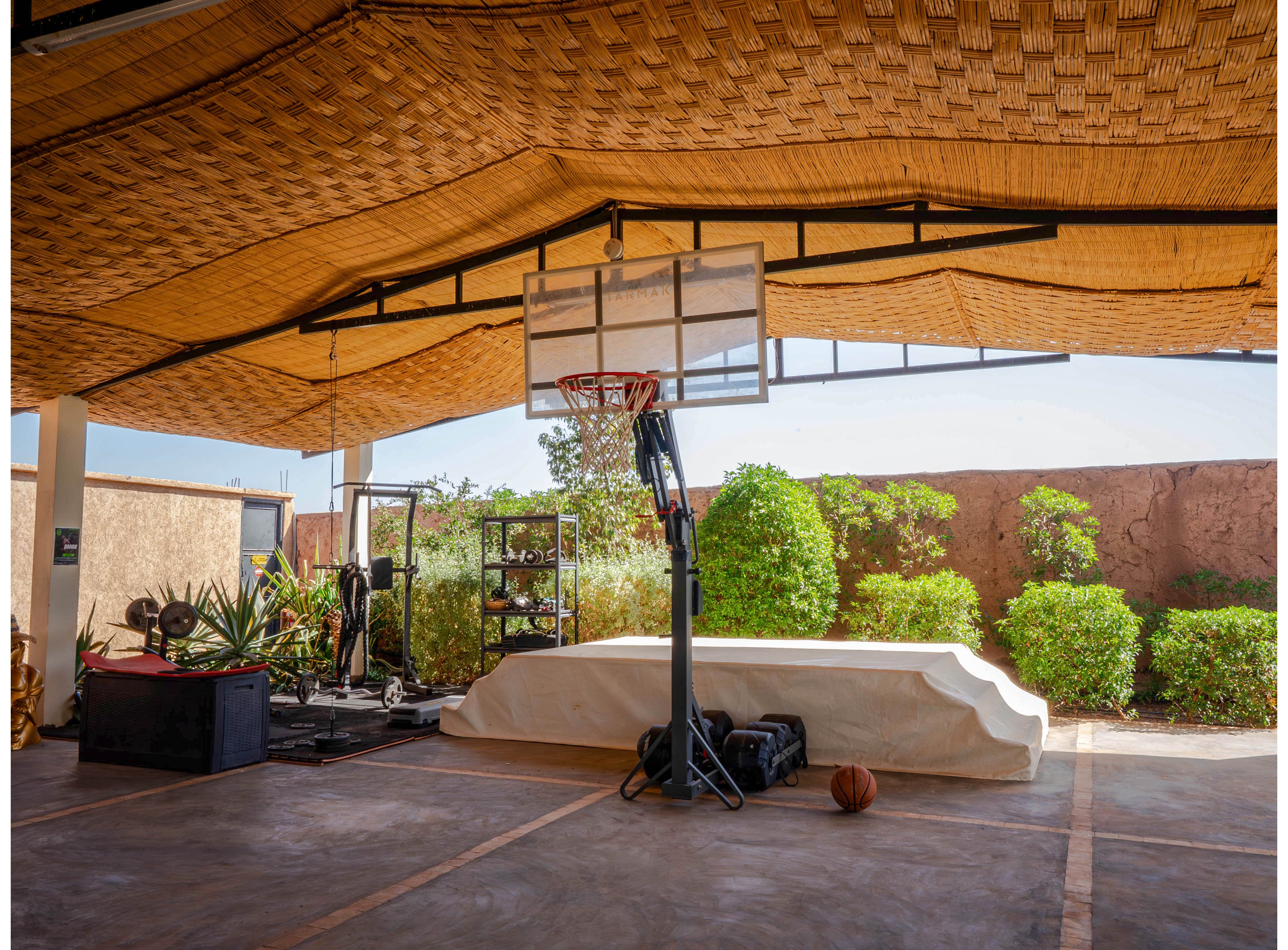 An adjustable basketball hoop stands under a woven canopy in an open-air courtyard. A single basketball rests on the ground near the base of the hoop, with sports bags and equipment nearby. Lush green shrubs line the perimeter against a high wall, adding contrast to the warm stone floor. The scene is brightly lit, suggesting a sunny, relaxed outdoor sports setting.
