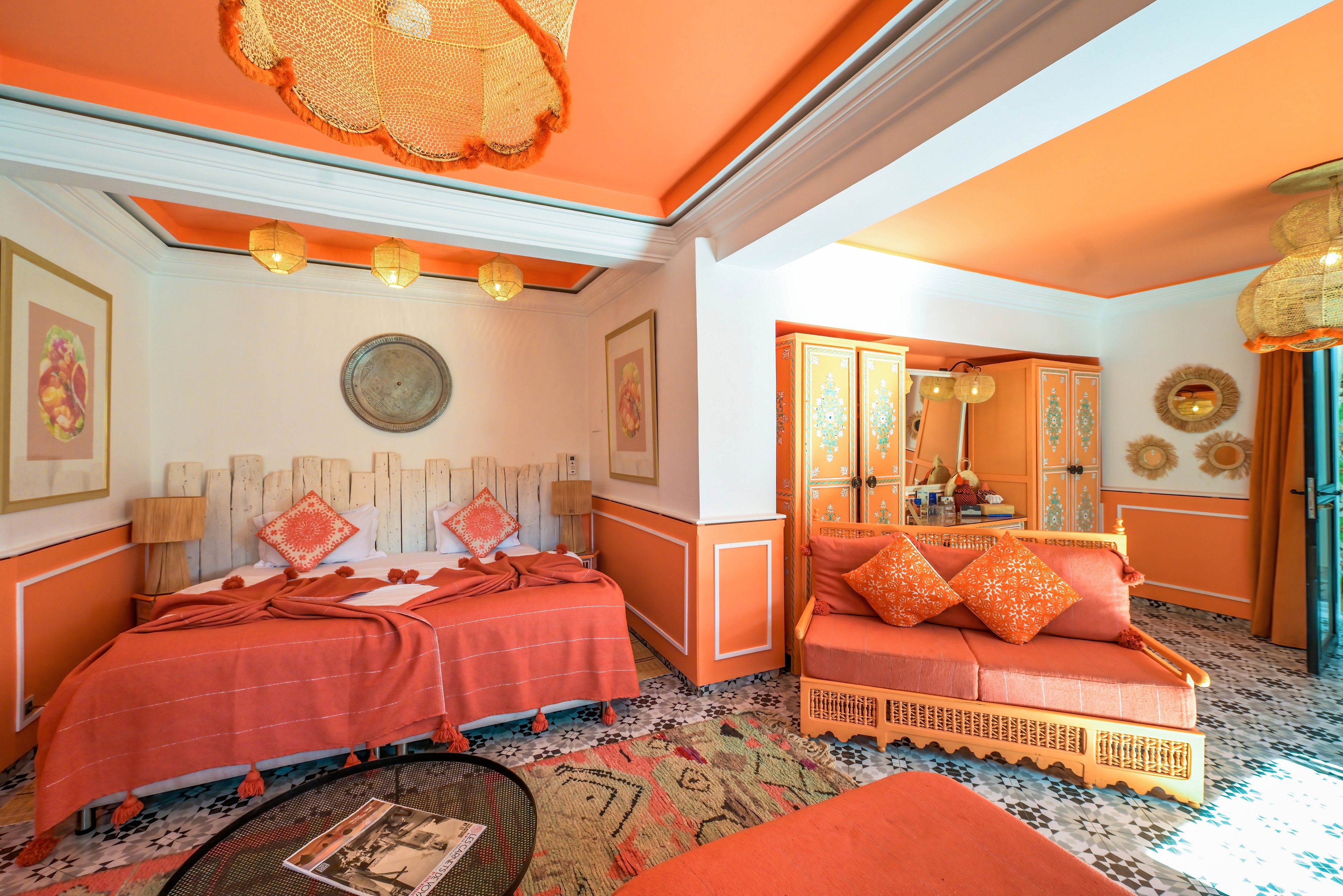 Vibrant hotel room interior featuring twin beds and a cozy sitting area in warm orange tones. The space includes patterned cushions, woven rattan furniture, and decorative wall art. Sunlight streams in, highlighting the tiled floor and eclectic decor. A magazine lies on a glass coffee table, adding a casual, lived-in feel to the scene.