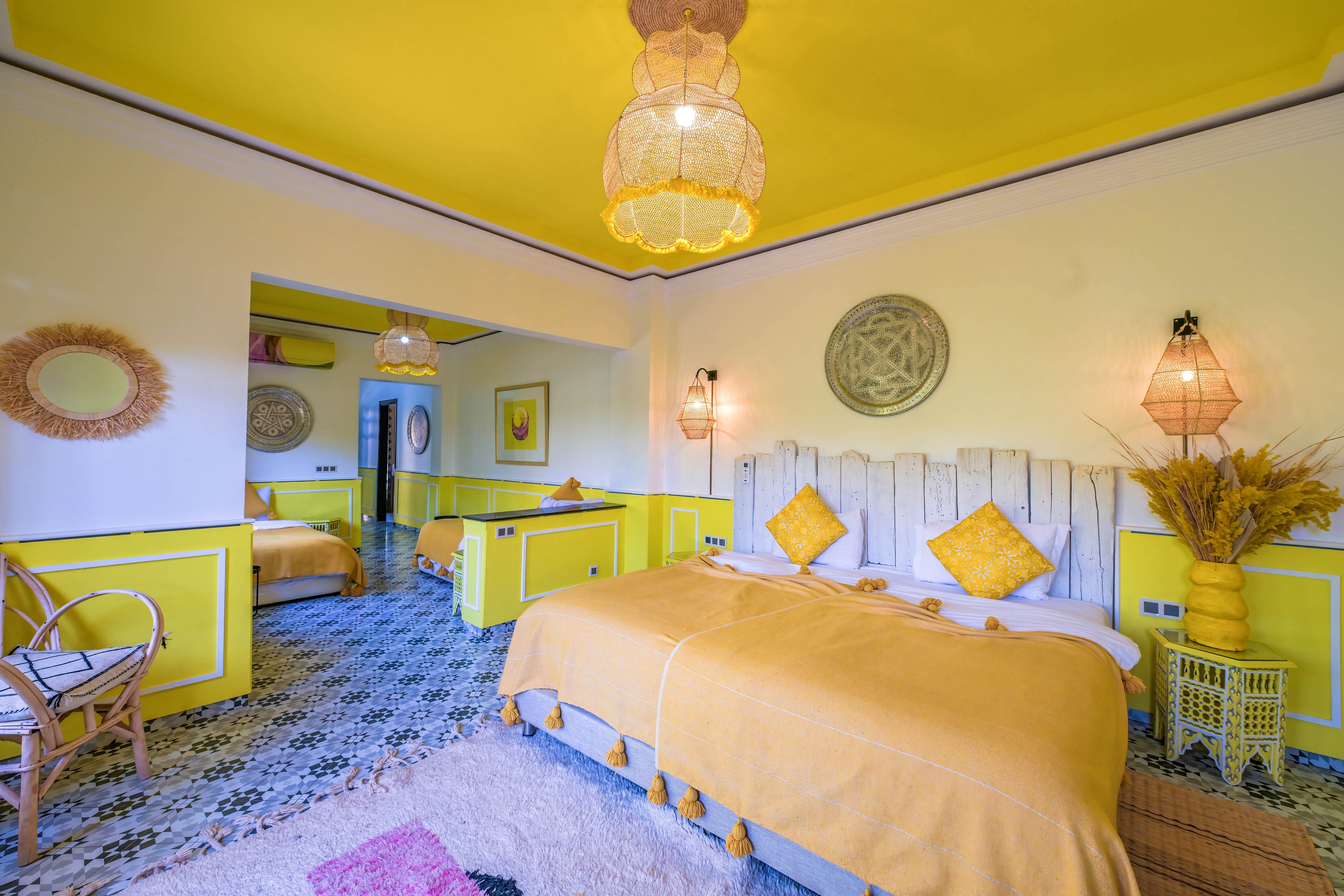 Colorful guest room with a large bed dressed in yellow bedding and matching pillows. The interior features bright yellow wall panels, a yellow ceiling, and patterned floor tiles. Wicker lamps, decorative mirrors, and dried flowers add a bohemian touch. An adjoining room with another bed is visible through an open doorway, enhancing the spacious feel.