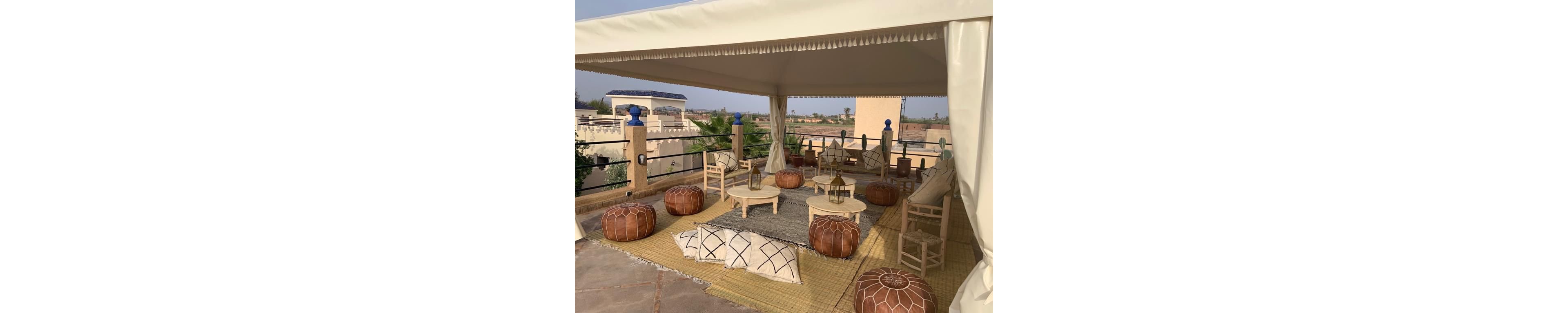 An outdoor lounge area is arranged beneath a large cream-colored canopy. Low wooden tables, leather poufs, and woven rugs create a cozy Moroccan-inspired seating space. Decorative lanterns sit on the tables and floor, surrounded by cushions with geometric patterns. The terrace overlooks distant buildings and greenery under a warm, hazy sky.