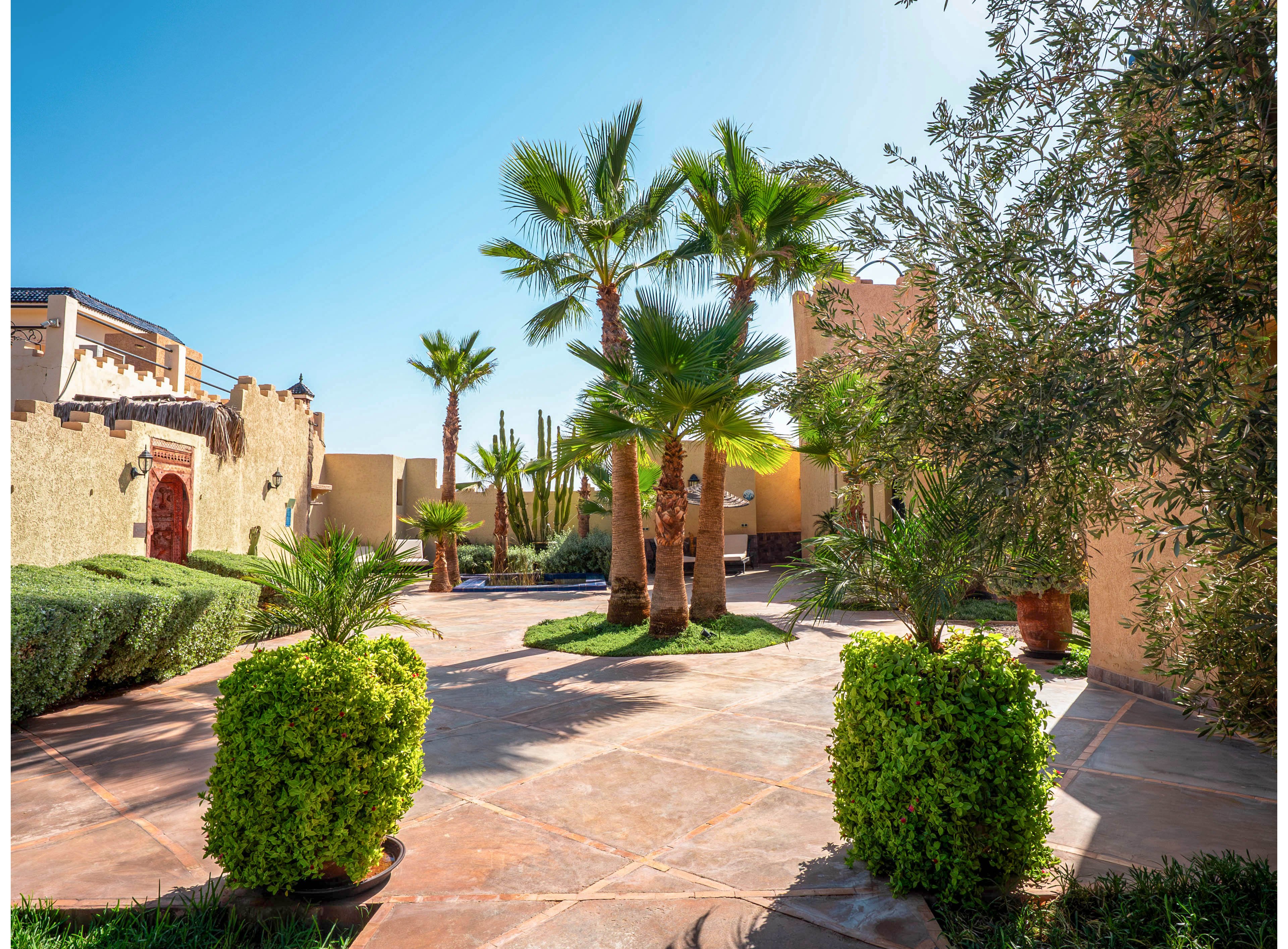 A bright courtyard features several tall palm trees surrounded by manicured shrubs and greenery. Warm-toned stone paving leads toward a villa-style building with textured walls and arched wooden doors. The clear blue sky and strong sunlight create sharp shadows and a vibrant, inviting atmosphere. The scene suggests a tranquil, resort-like outdoor space in a warm climate.