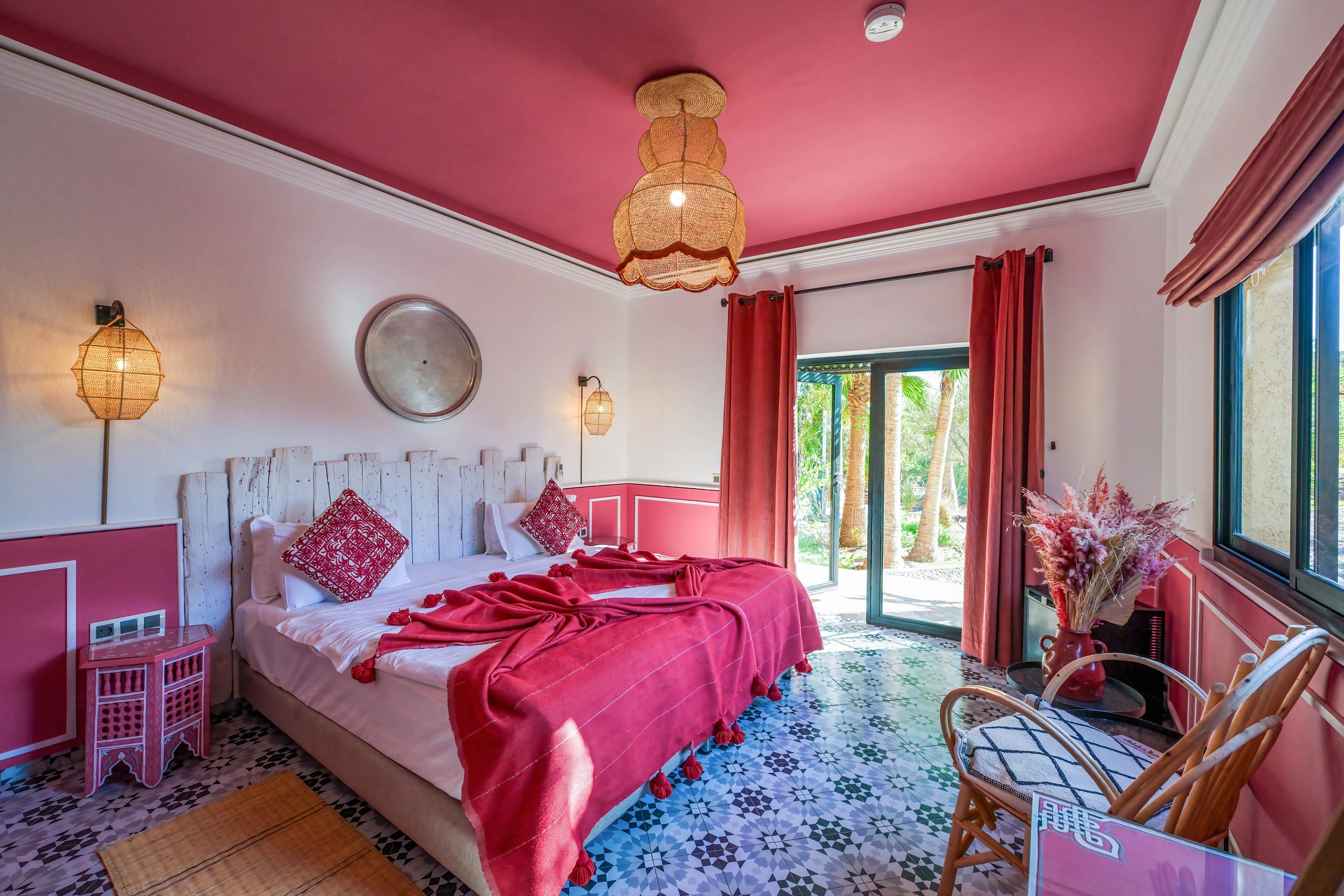 A stylish boutique bedroom features a double bed with red bedding and patterned pillows. The room is decorated in shades of red and pink, with a matching ceiling, curtains, and accent furniture. Natural light enters through large glass doors and a side window, revealing greenery outside. Patterned floor tiles, woven lighting fixtures, and dried floral arrangements add a cozy, eclectic atmosphere.
