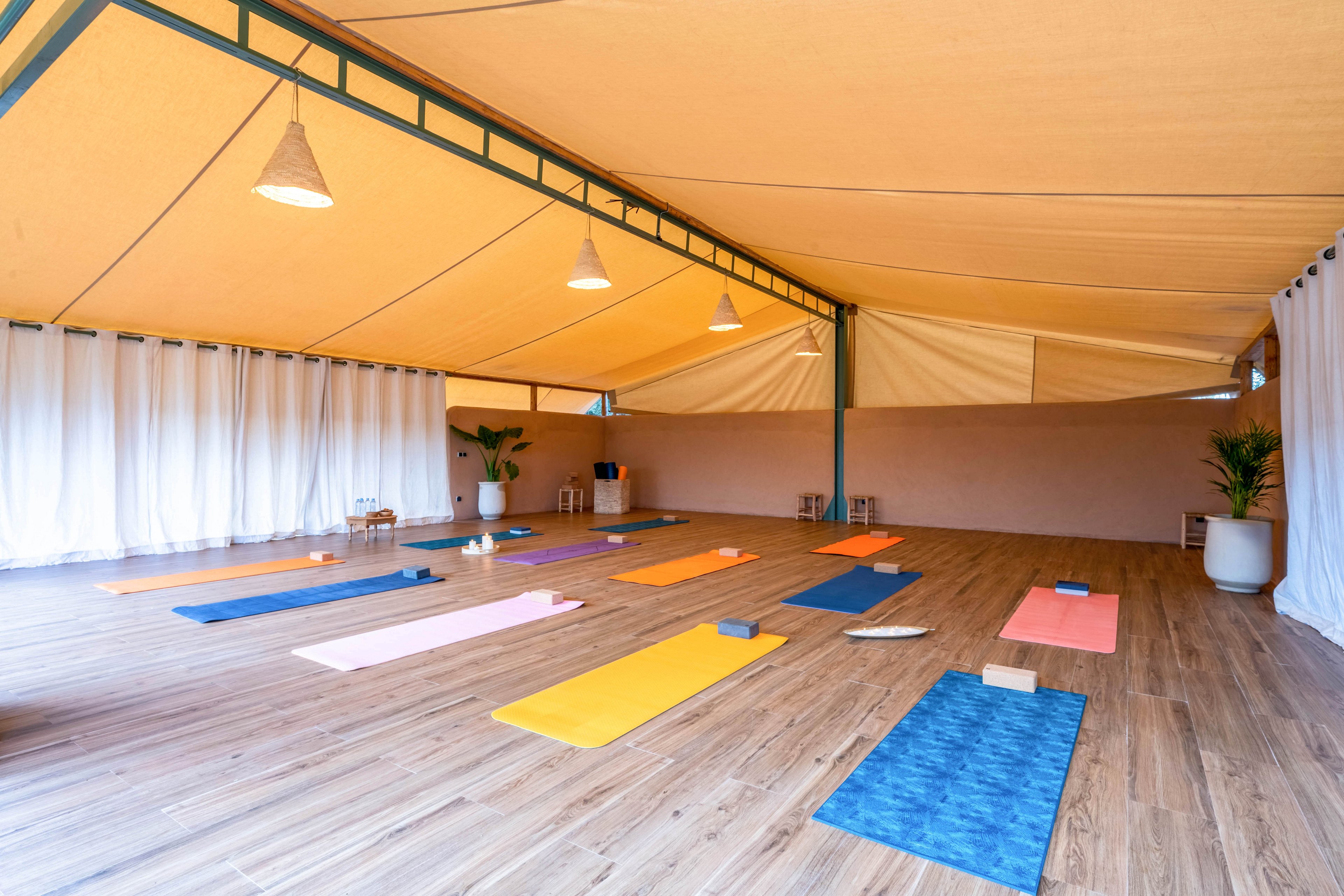 An open, covered yoga studio features neatly arranged colorful mats on a wooden floor. The space is bright and airy, with a large fabric canopy ceiling and soft hanging lights. Yoga blocks and props are placed beside several mats, suggesting preparation for a class. White curtains and potted plants along the walls create a calm, minimalist atmosphere.