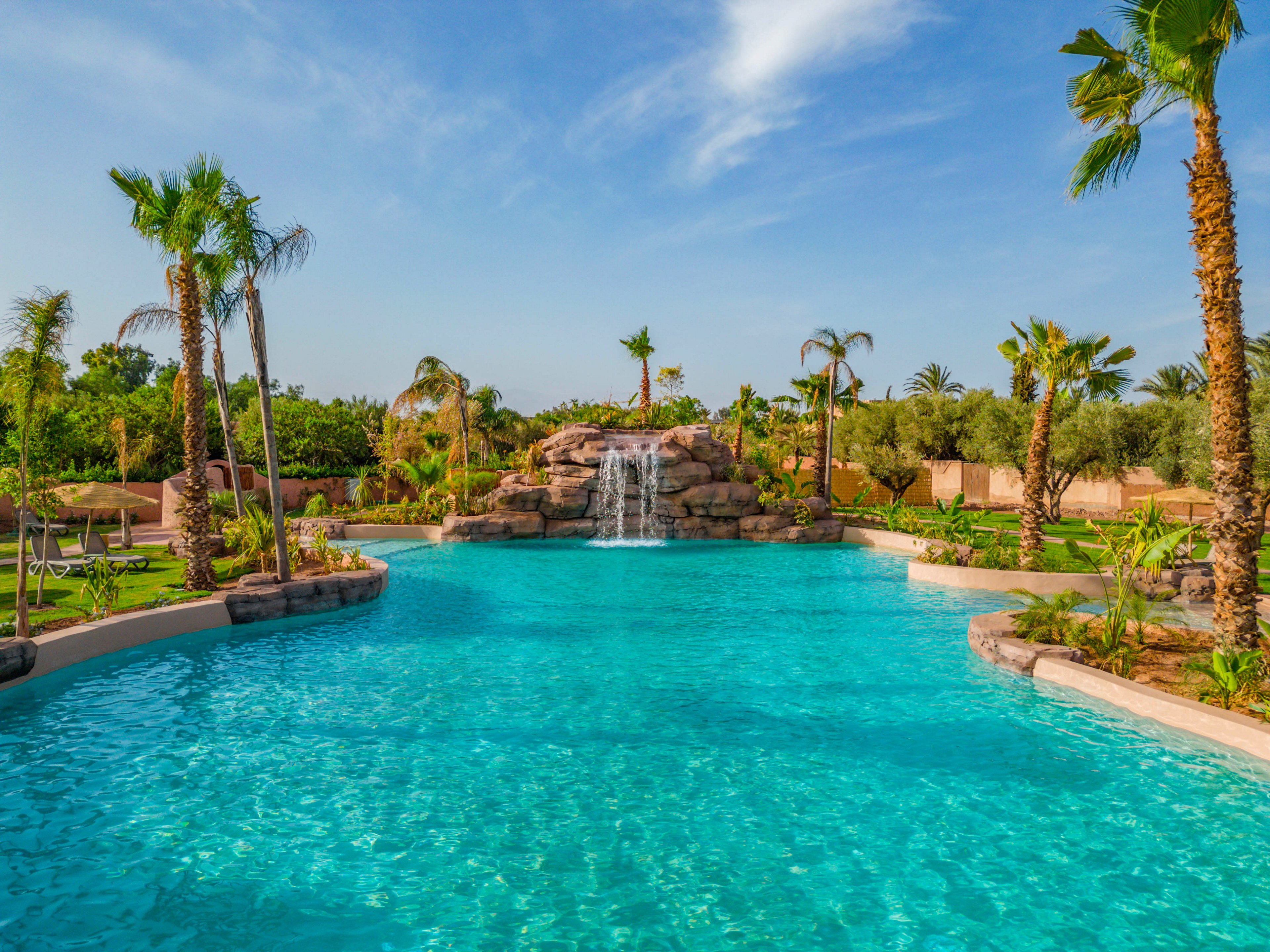 A large turquoise swimming pool stretches out under a bright blue sky. At the far end, a rock formation features a cascading waterfall flowing into the pool. Palm trees and lush green landscaping surround the water, creating a tropical resort atmosphere. The scene is calm and inviting, suggesting a relaxing vacation setting.