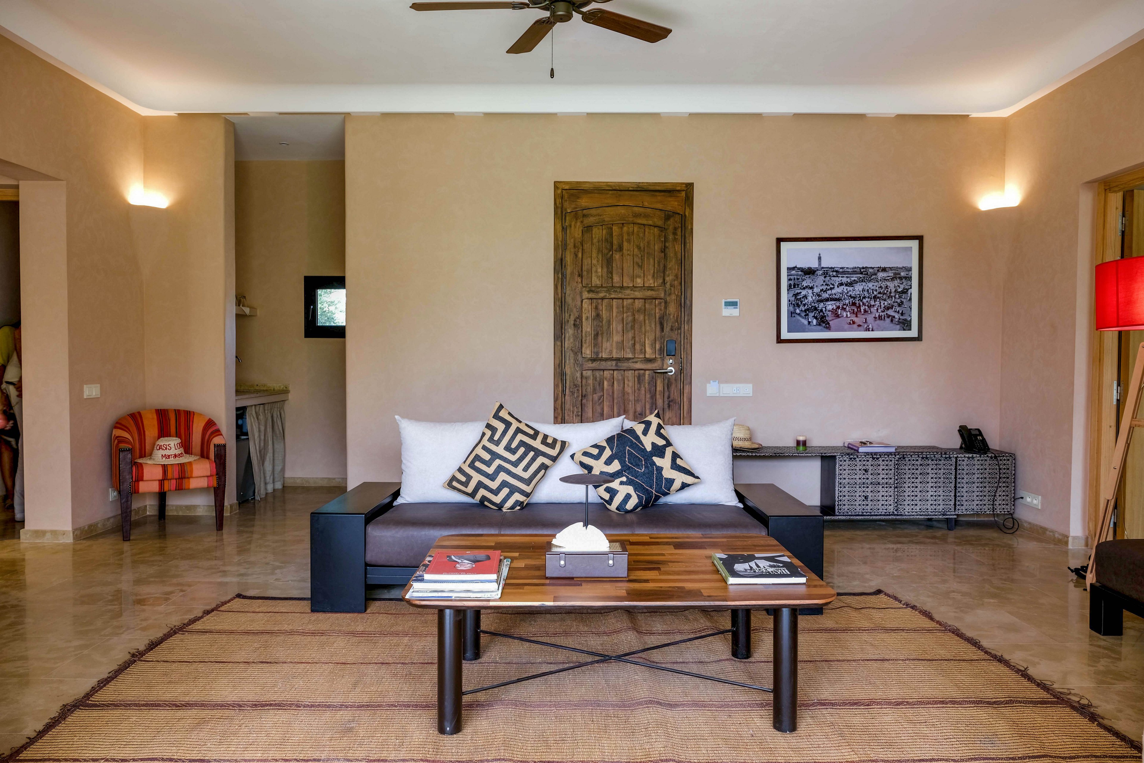 A contemporary living room features a central wooden door and a sleek sofa with patterned throw pillows. A low wooden coffee table holds books and a tissue box, placed on a large woven rug. Warm wall sconces, a ceiling fan, and neutral-colored walls create a cozy atmosphere. Additional seating, framed artwork, and a red floor lamp add subtle accents to the modern interior.