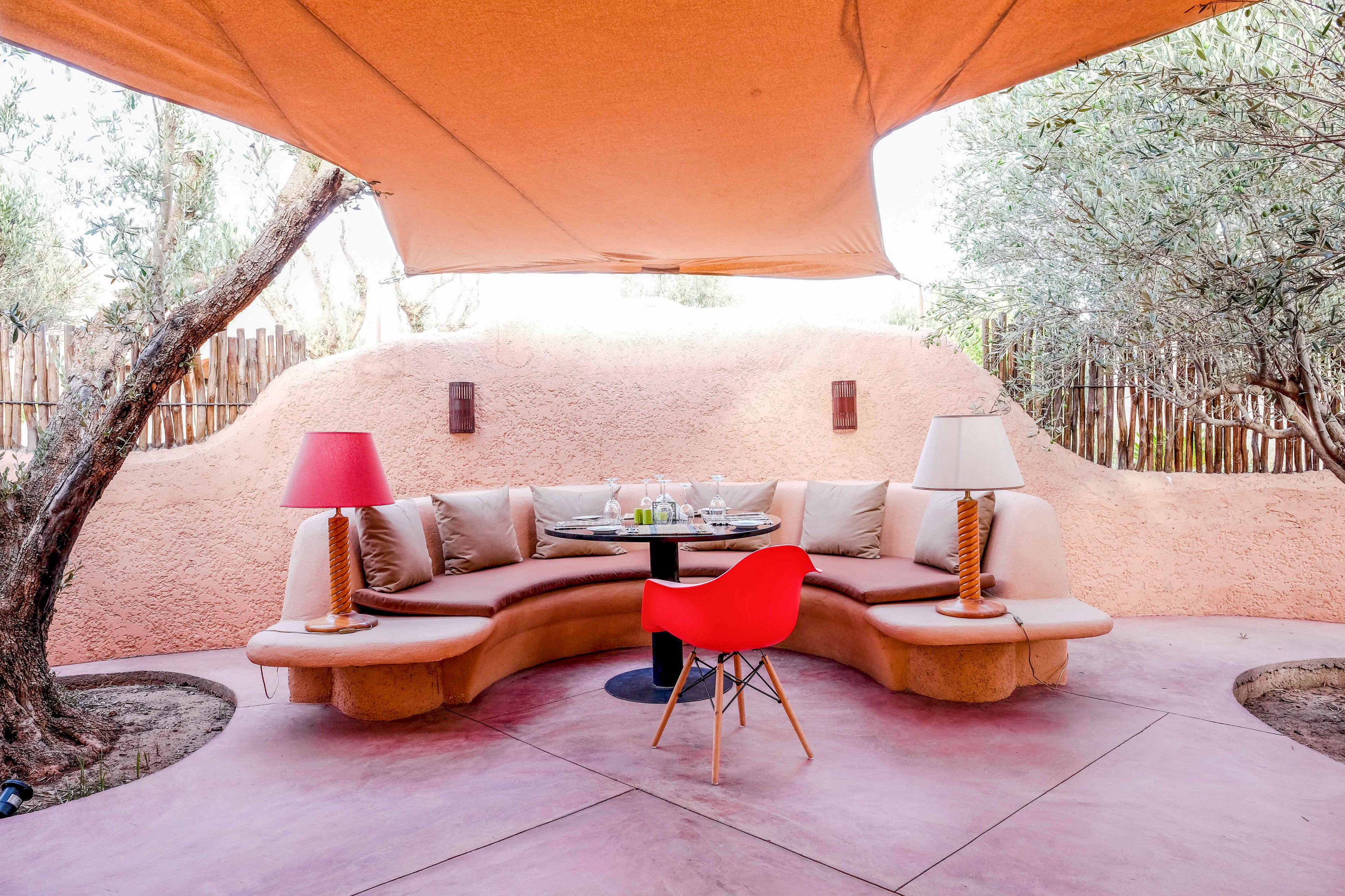 A stylish outdoor seating area features a curved built-in sofa with neutral cushions arranged around a small round table. The space is shaded by a large fabric canopy and framed by organic stucco walls and wooden fencing. Two table lamps with rope-wrapped bases and a bold red chair add color accents to the warm-toned patio. Trees grow through the floor, enhancing the natural, relaxed atmosphere of the lounge.