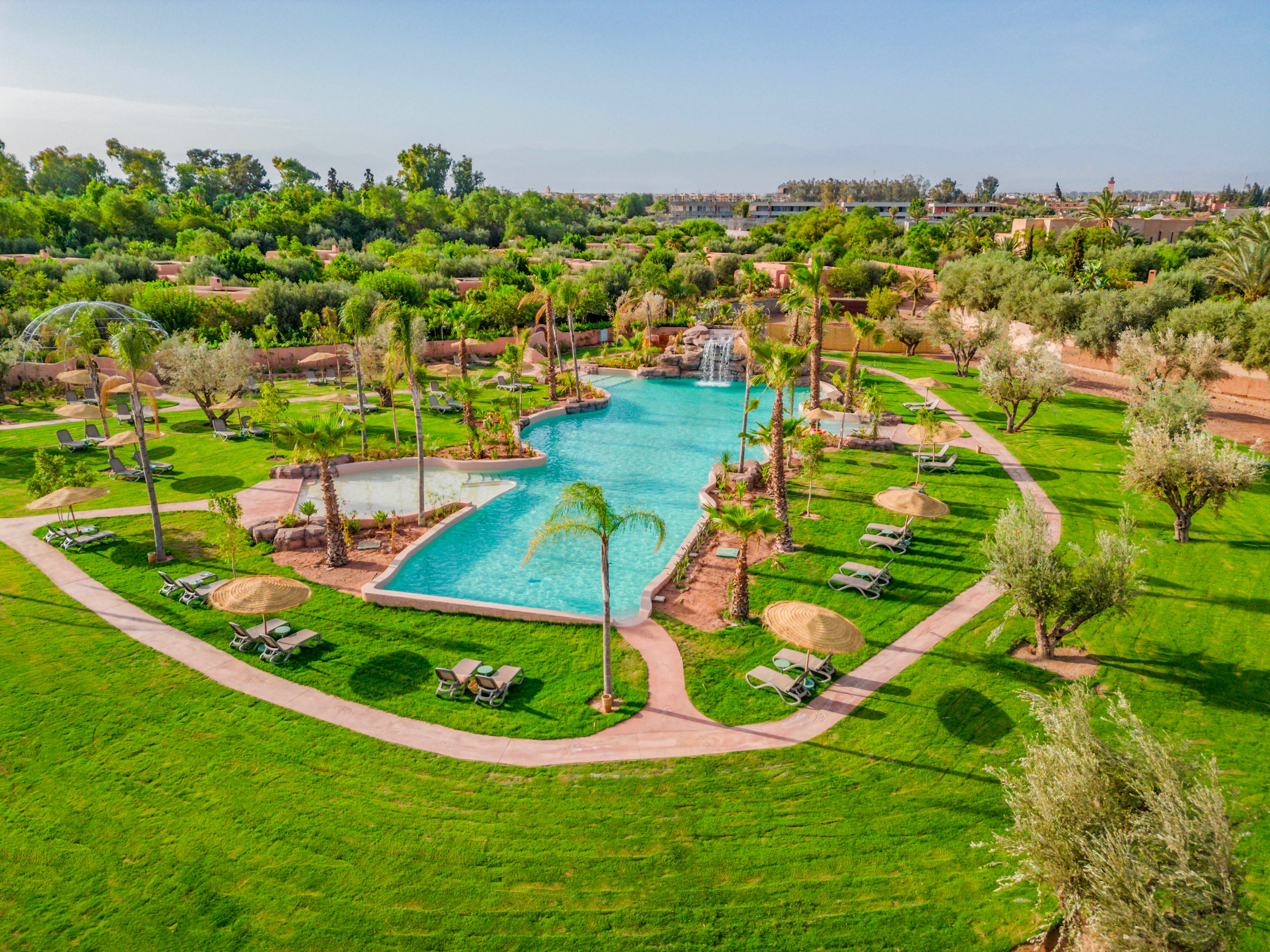 An aerial view showcases a large, lagoon-style swimming pool surrounded by lush green lawns and palm trees. The pool features a small waterfall and rock formations, creating a tropical oasis atmosphere. Sun loungers and umbrellas are arranged neatly around the water, suggesting a relaxing resort setting. In the background, gardens and trees extend toward the horizon under a clear blue sky.