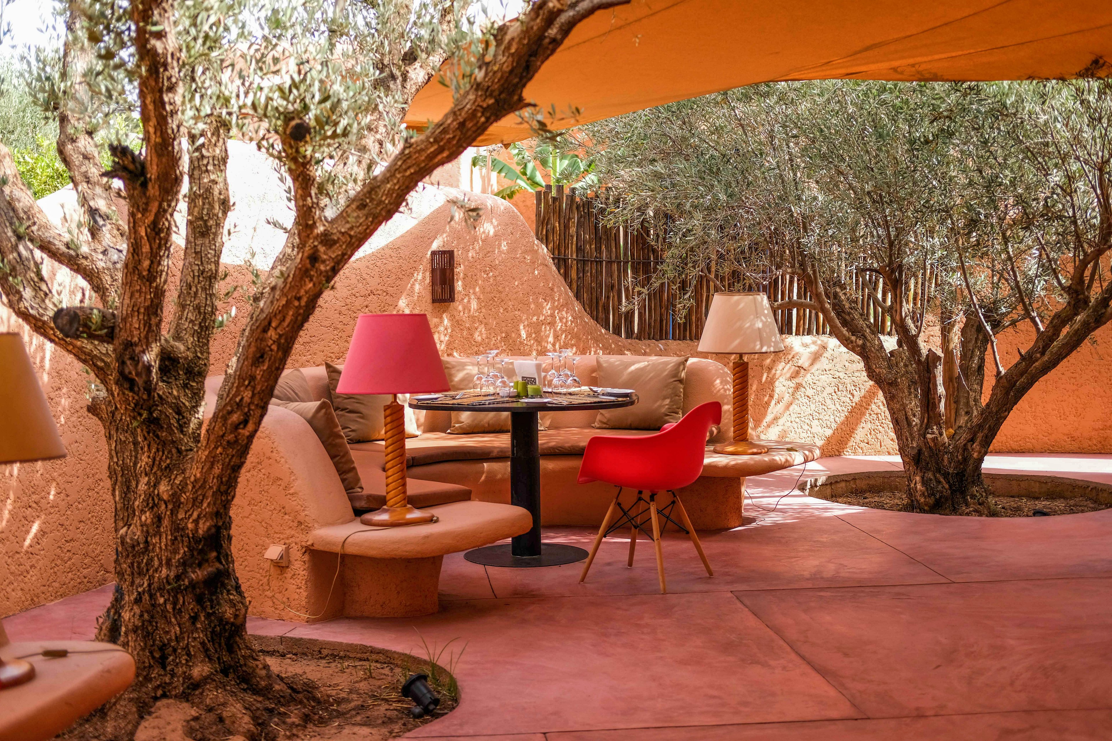 An inviting outdoor seating area is arranged beneath mature trees, creating a shaded patio space. Curved benches with cushions surround a round dining table set with glassware and plates. Warm-toned walls and flooring in terracotta hues complement the soft lighting from table lamps. A bright red chair adds a pop of color against the earthy surroundings, enhancing the relaxed, stylish atmosphere.