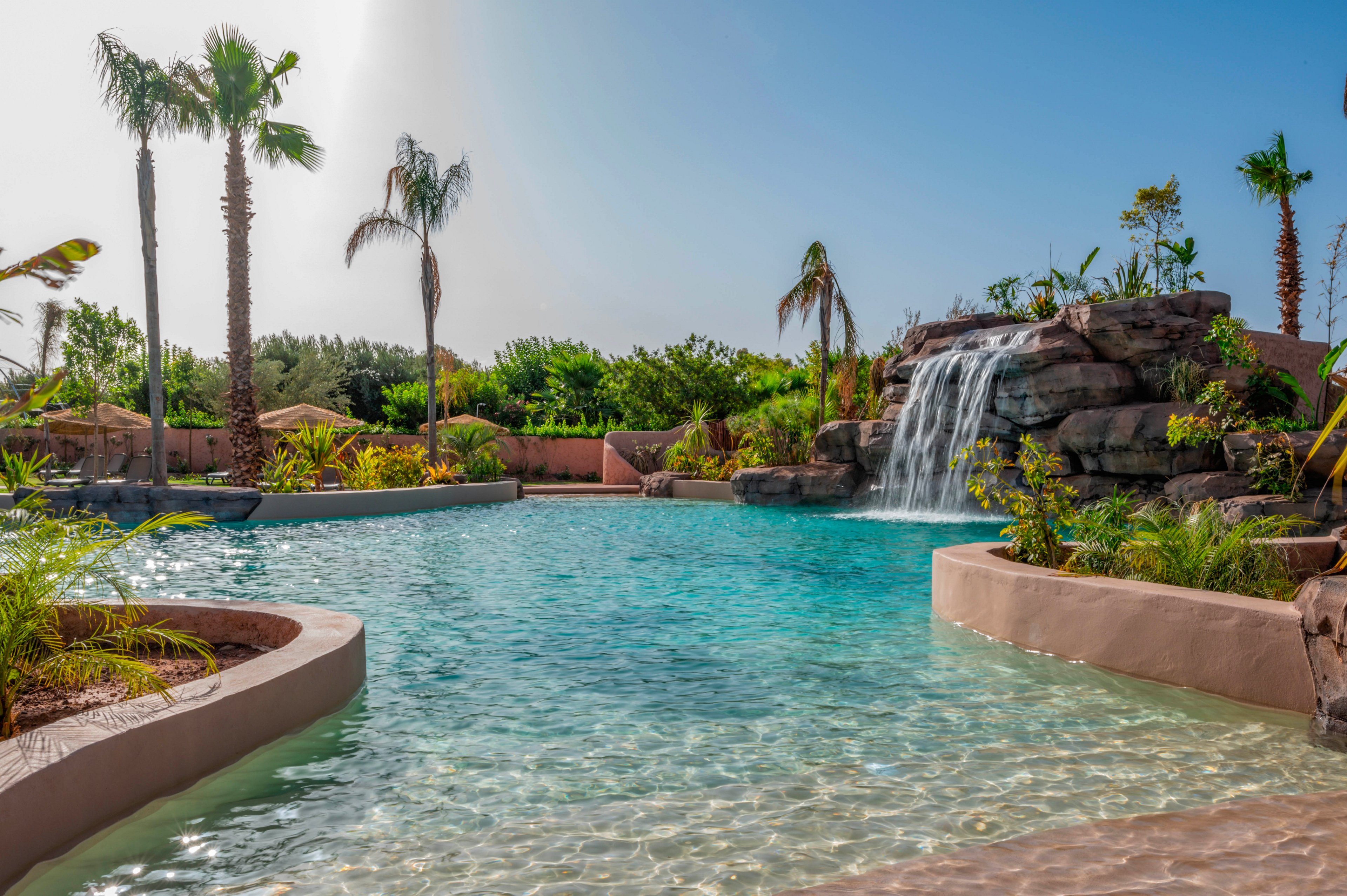 A large turquoise swimming pool features a rock waterfall cascading into the water. Palm trees and lush tropical plants surround the pool, creating a resort-like atmosphere. Curved concrete edges and shallow entry areas frame the clear blue water. The bright sunny sky and calm surface of the pool evoke a relaxing vacation setting.