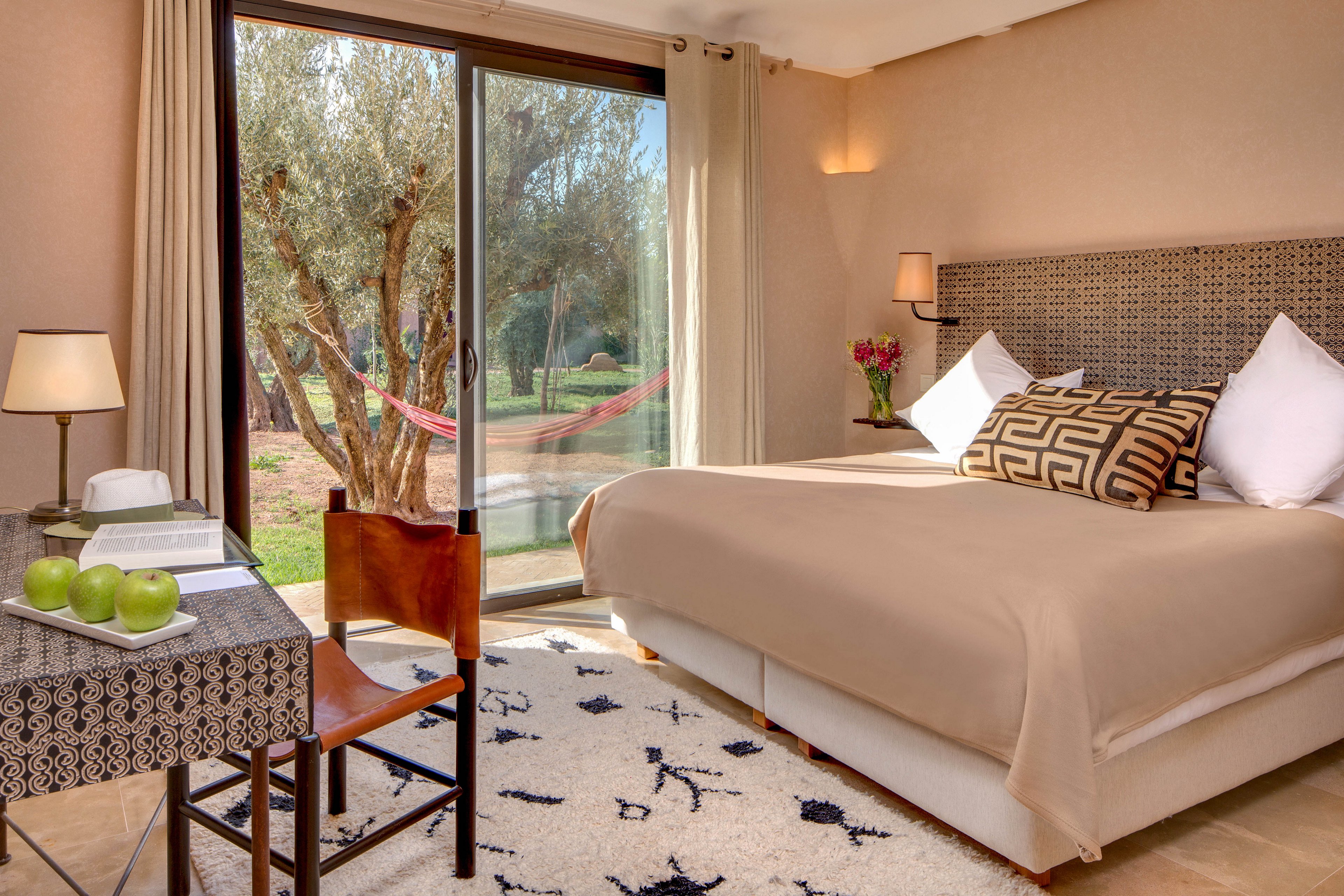 A neatly made double bed with neutral bedding dominates a bright hotel bedroom. Large sliding glass doors open to a garden with trees and a hammock outside. A small table with green apples, books, and a chair sits near the window, while a patterned rug and cushions add texture. Warm wall lighting and beige curtains create a cozy, inviting atmosphere.