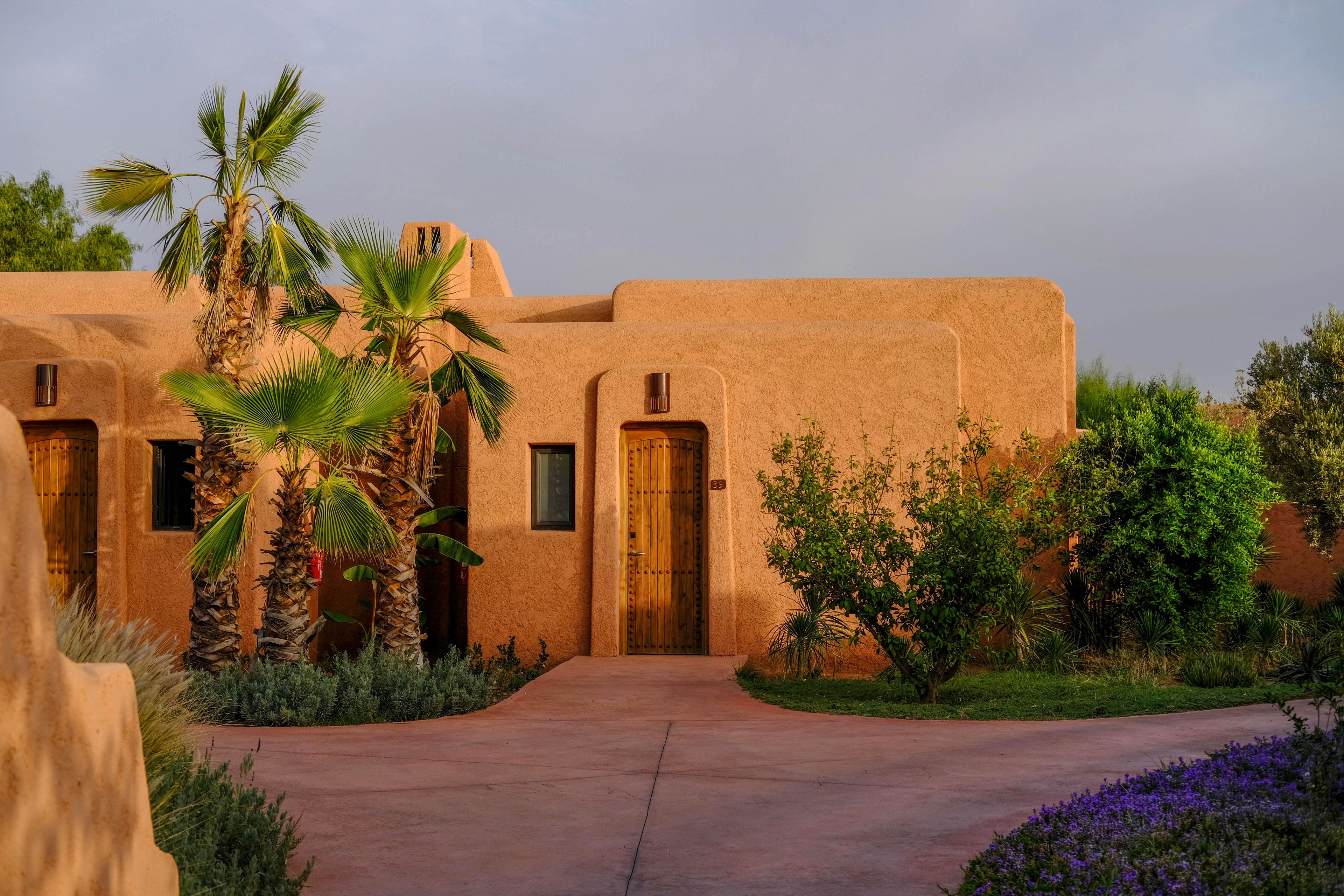 A warm-toned adobe-style villa stands under a soft sky, framed by lush greenery and palm trees. The building features simple rectangular forms and wooden doors that open onto a smooth, curved pathway. Late afternoon or early evening light casts gentle shadows, enhancing the earthy colors of the walls and surrounding plants. Purple flowers and manicured shrubs add contrast and a tranquil, resort-like atmosphere.