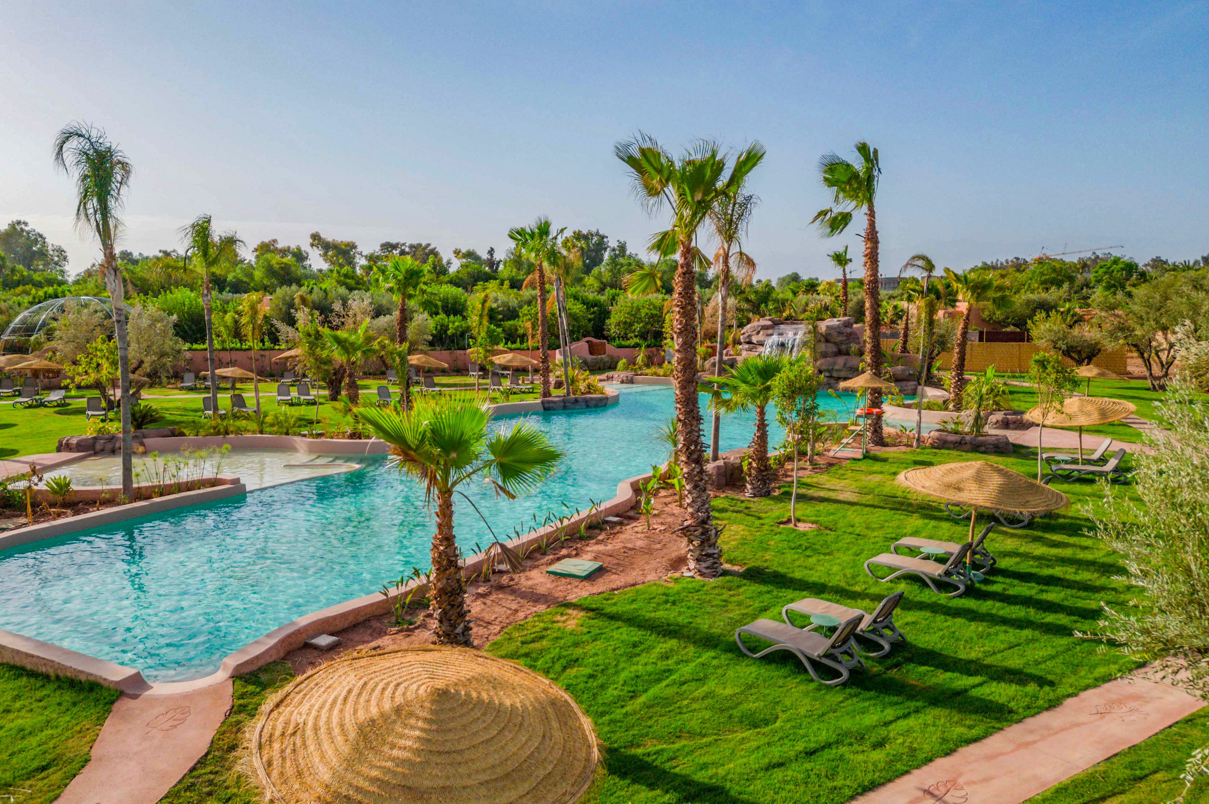 A large resort-style swimming pool winds through a lush tropical garden filled with palm trees and manicured lawns. Sun loungers and straw parasols are arranged neatly along the poolside paths, suggesting a relaxing vacation setting. The clear blue water contrasts with the vibrant green grass and foliage under a bright, cloudless sky. The scene feels tranquil and inviting, ideal for leisure and sunbathing.