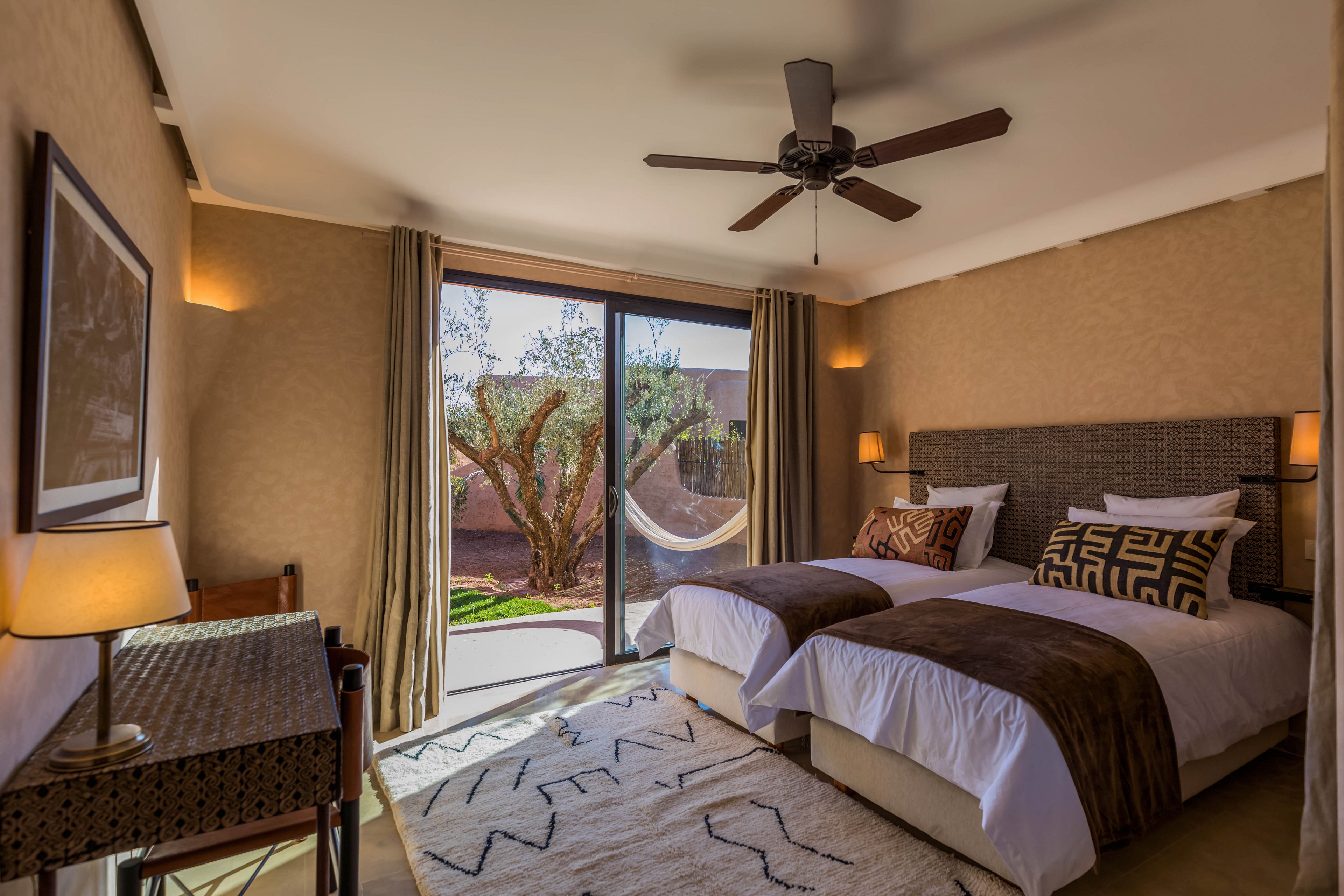 A warm, contemporary bedroom features two twin beds with brown throws and patterned cushions. Large glass doors open to a garden with a tree and hammock, allowing natural light to fill the room. A ceiling fan, bedside lamps, and a desk with a lamp create a comfortable and functional space. Neutral walls and a textured rug contribute to the cozy, inviting atmosphere.