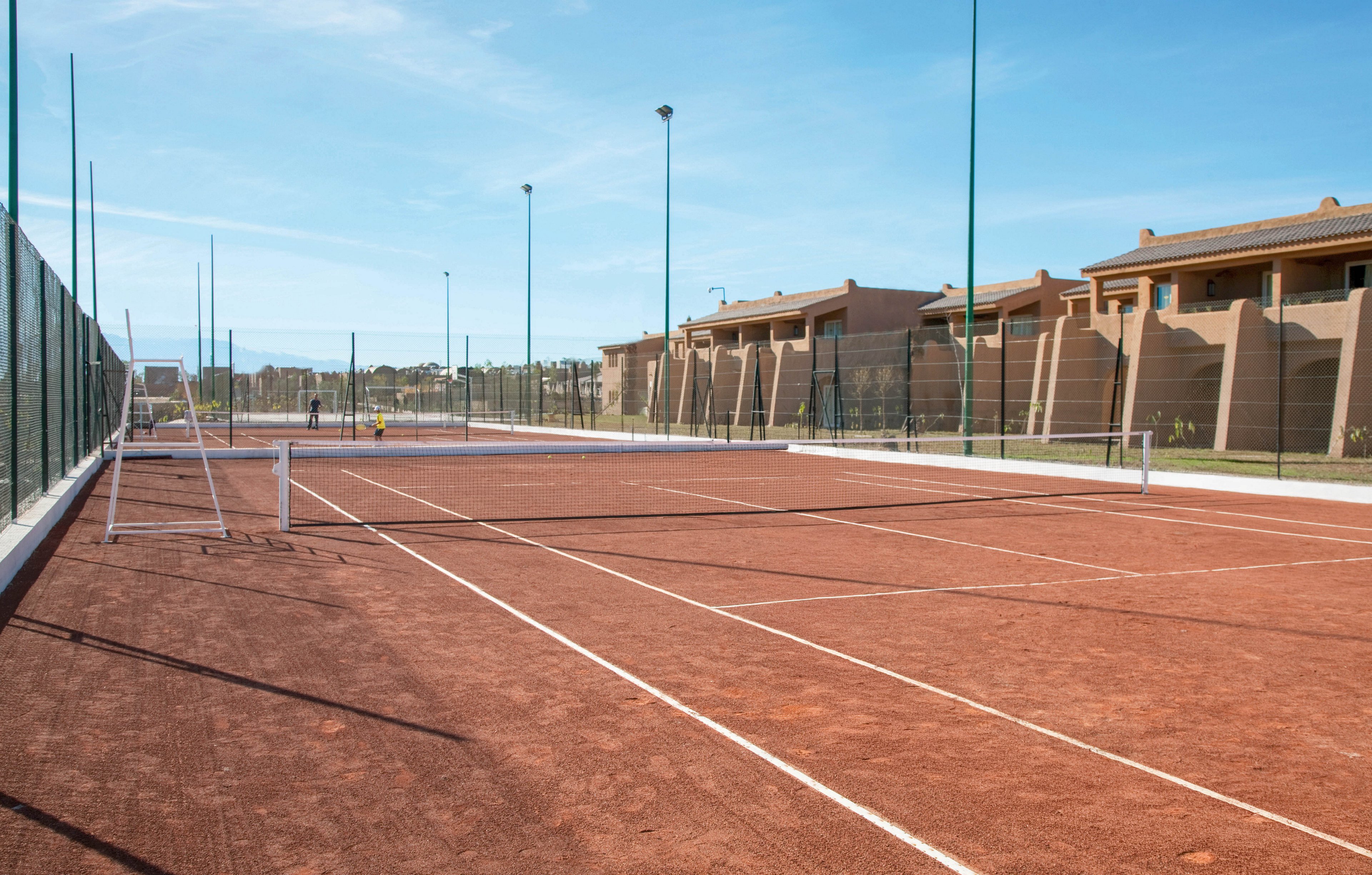 Four people are engaged in a doubles tennis game on a clay court, with a referee seated on a high chair. The setting is outdoors, surrounded by residential buildings and fencing, under a clear blue sky. The players appear to be adults, both male and female, dressed in athletic attire. The scene conveys an energetic and competitive mood.