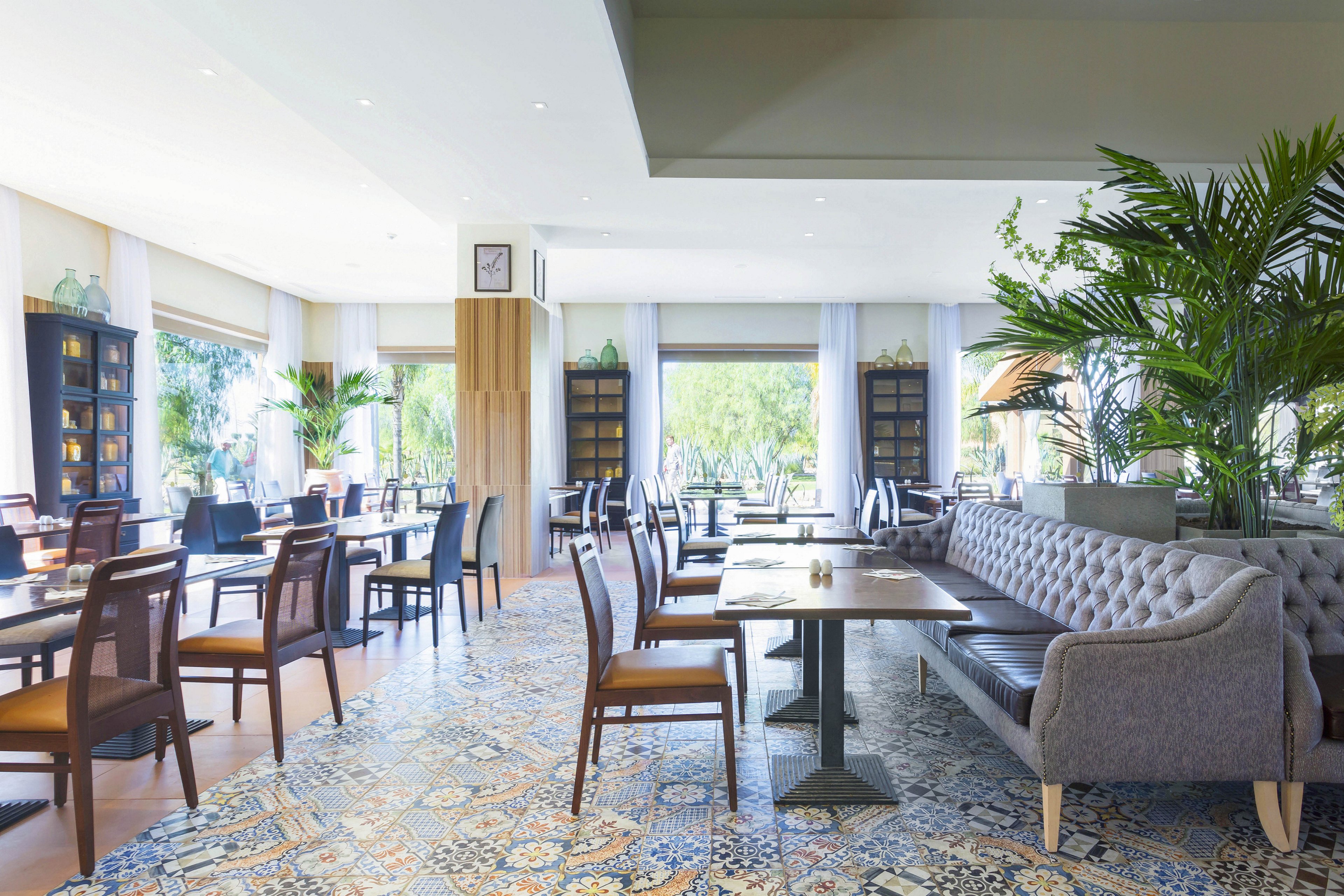 The image shows a spacious restaurant dining area featuring elegant wooden chairs and a tufted sofa bench. Large windows allow natural light to flood the space, highlighting decorative patterned floor tiles and lush green plants. The setting is airy and contemporary, with neutral tones and a relaxed atmosphere. A wall clock is visible above a wooden column, adding a subtle numeric element.