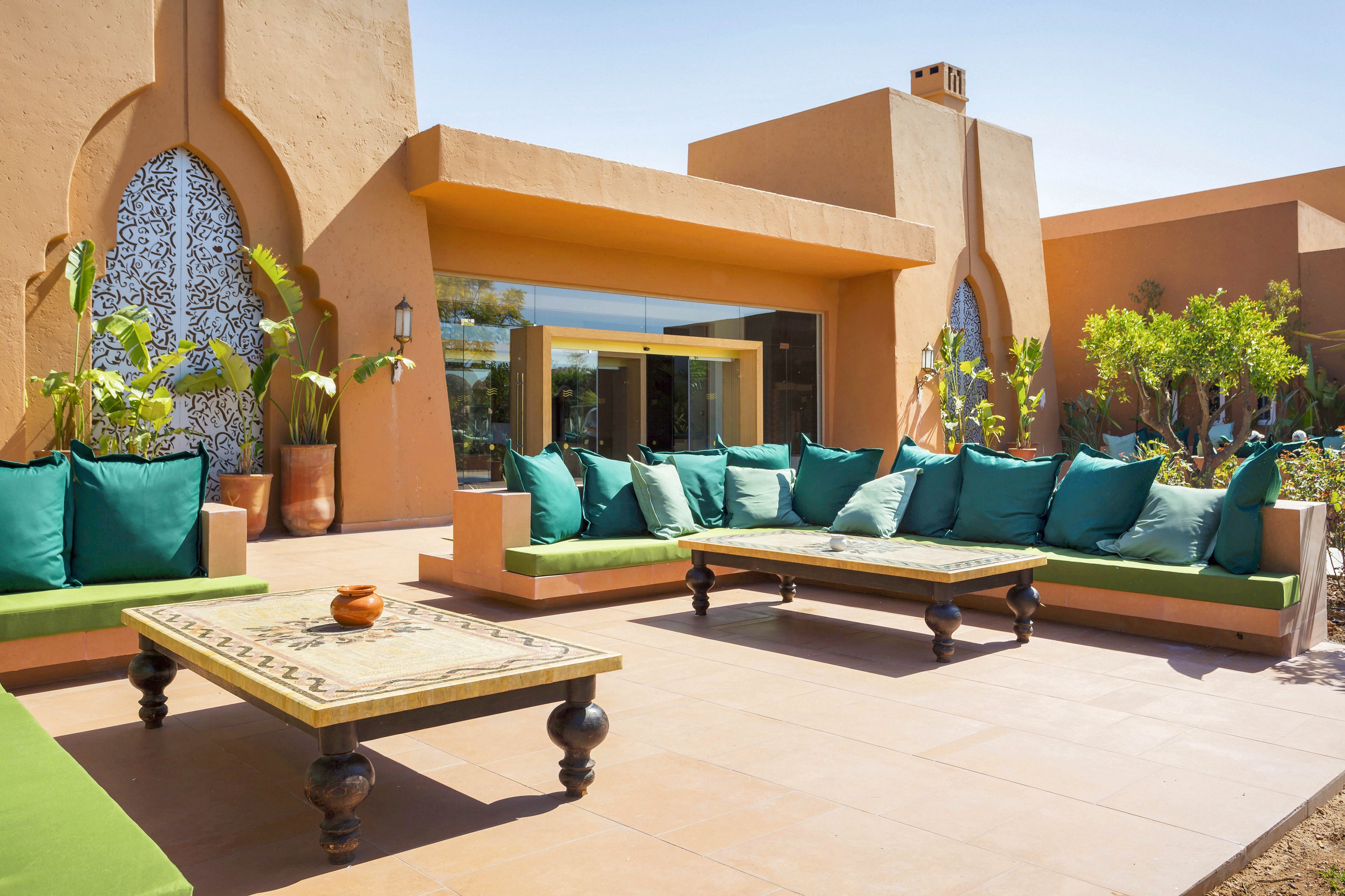 The image features a spacious outdoor seating area with long sofas adorned with green and teal cushions. The setting is a sunny patio with terracotta tiles, surrounded by lush plants and contemporary architecture. Two wooden tables with decorative legs are placed in front of the sofas, and the overall mood is relaxed and inviting. No visible text or numeric values are present.