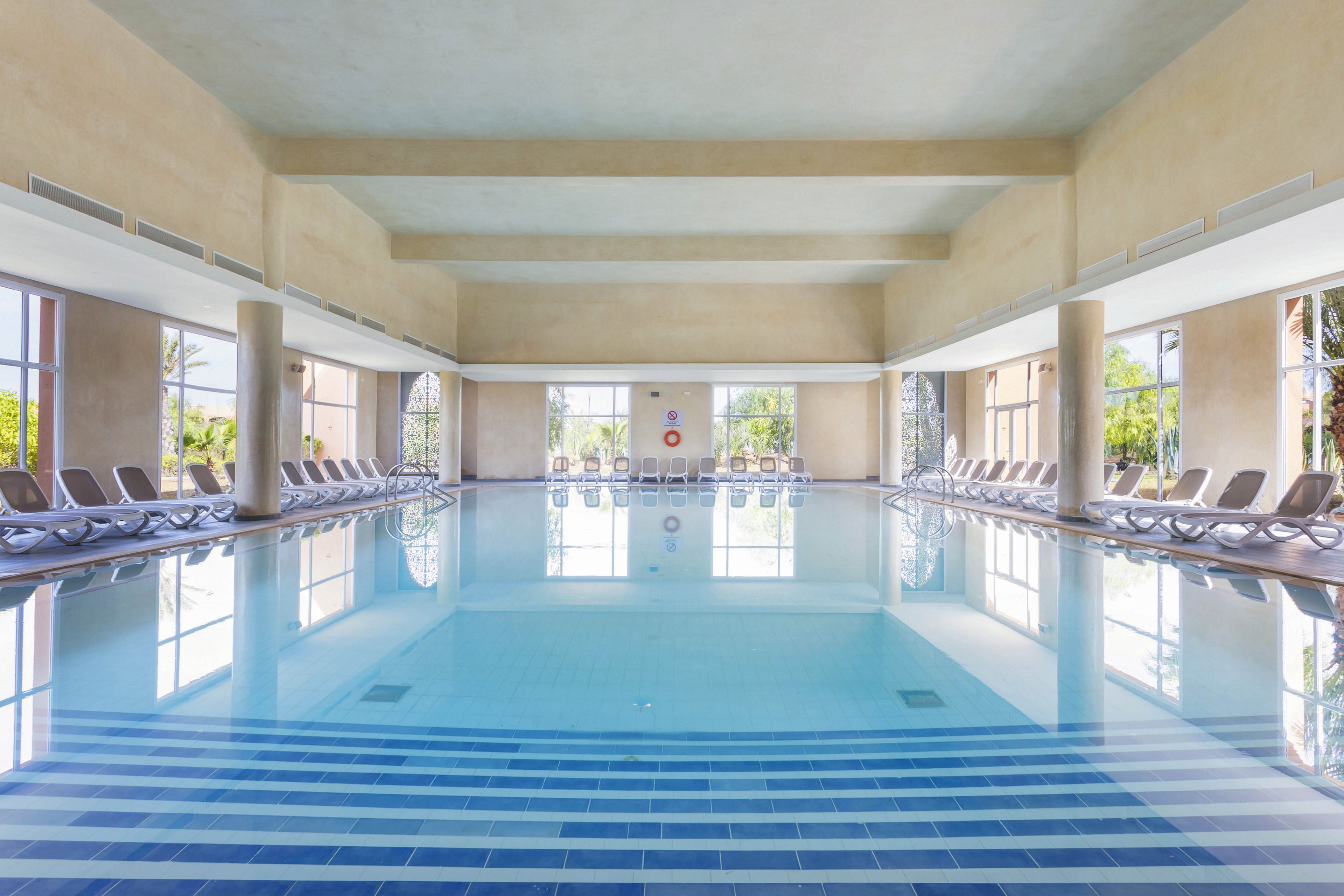 A large, serene indoor swimming pool is the main focus, surrounded by rows of empty lounge chairs. The pool area features high ceilings and large windows, allowing natural light to fill the space and offering views of greenery outside. The atmosphere is calm and inviting, with a clean, modern design and soft neutral tones. A red lifebuoy is visible on the far wall, adding a pop of color.