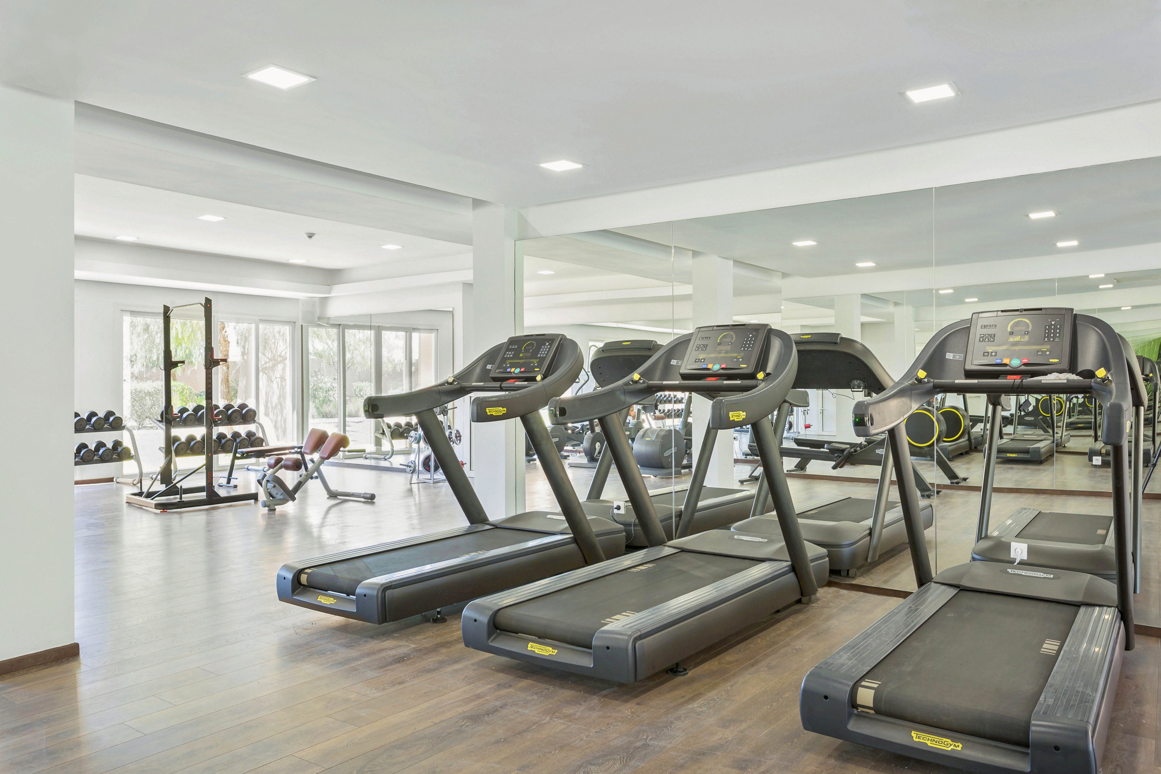 The image shows a spacious fitness center featuring three treadmills prominently in the foreground. In the background, there are free weights and benches near large windows that let in natural light. The setting is clean and contemporary, with light walls and wood flooring. No visible people or text are present, and the mood is bright and inviting.