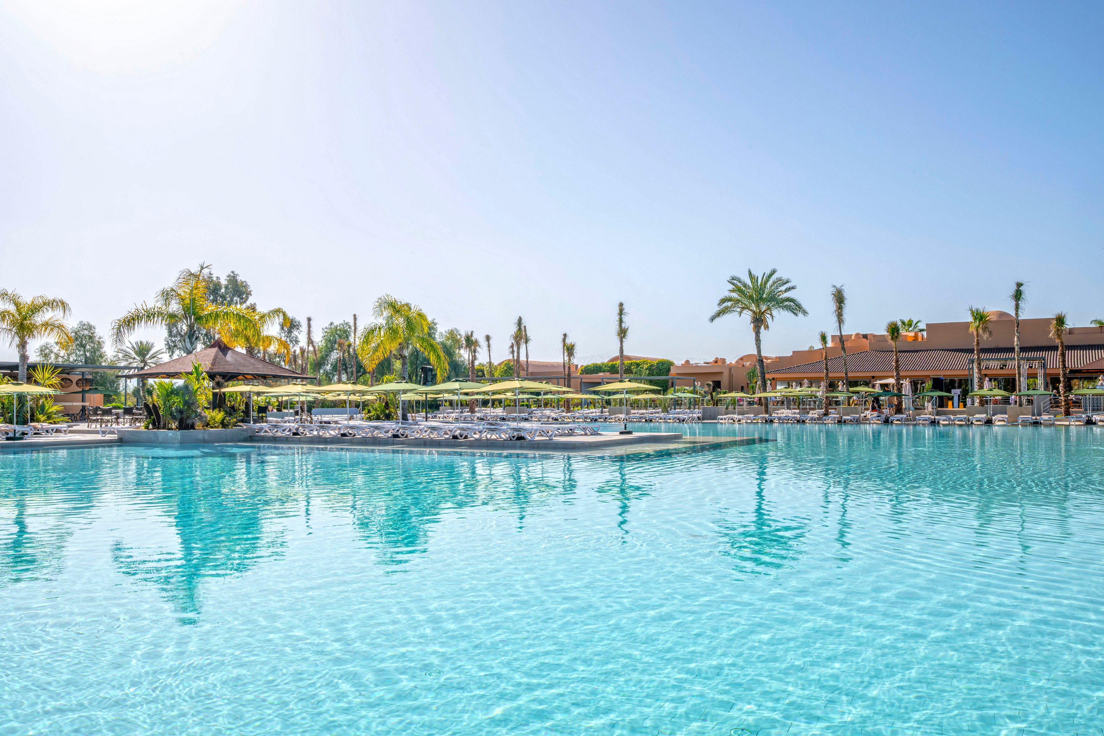 A large, clear blue swimming pool is the main focus, surrounded by palm trees and sun loungers. The setting appears to be a resort or hotel with low-rise buildings in the background under a bright, cloudless sky. The tranquil water and tropical landscaping create a relaxing vacation atmosphere.