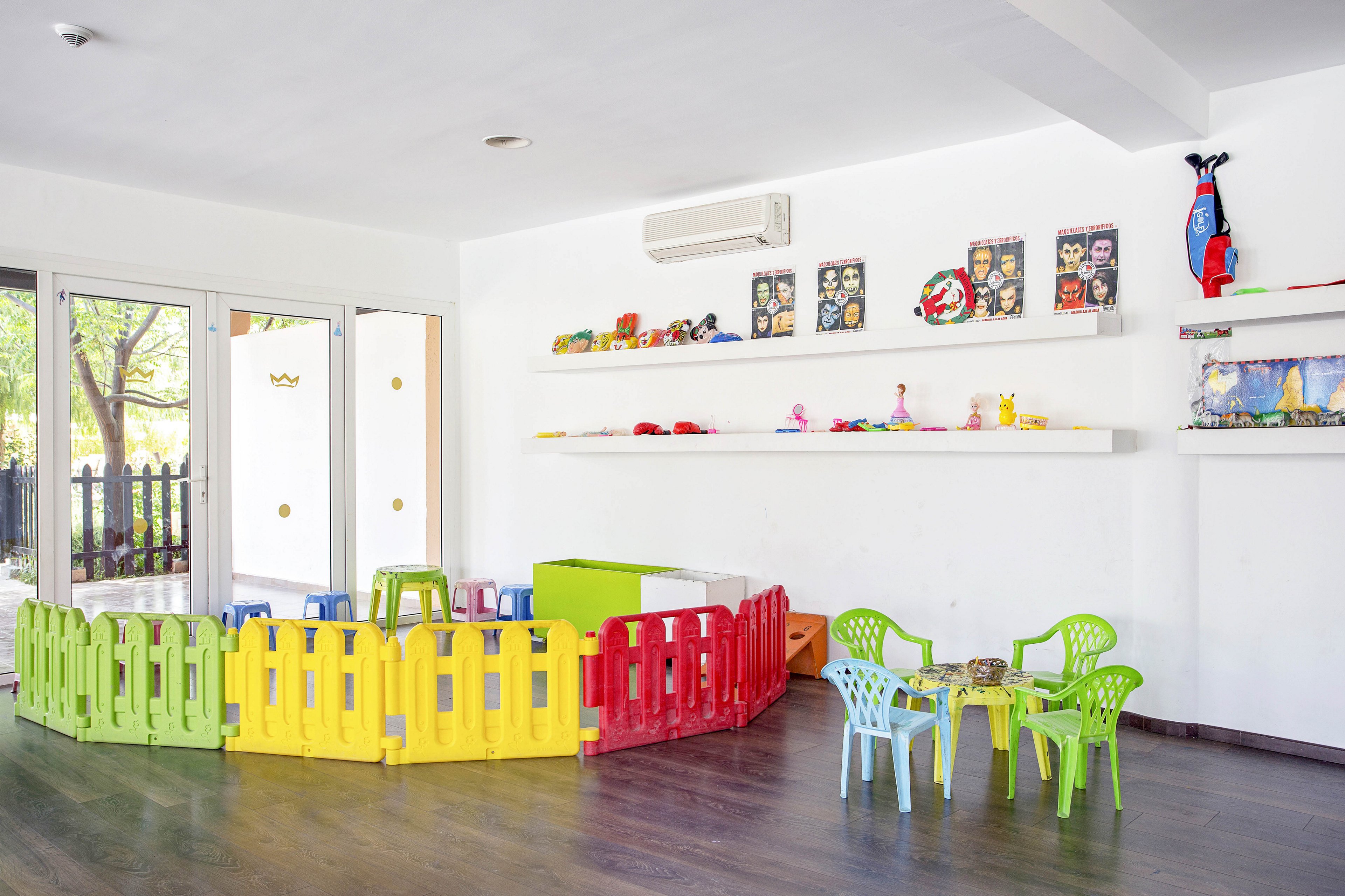 A spacious children's playroom features vibrant yellow and red plastic fences forming a safe play zone. Green and blue plastic chairs surround a small table, while shelves display toys and art supplies. Large windows let in natural light, creating a cheerful and inviting atmosphere for young children.