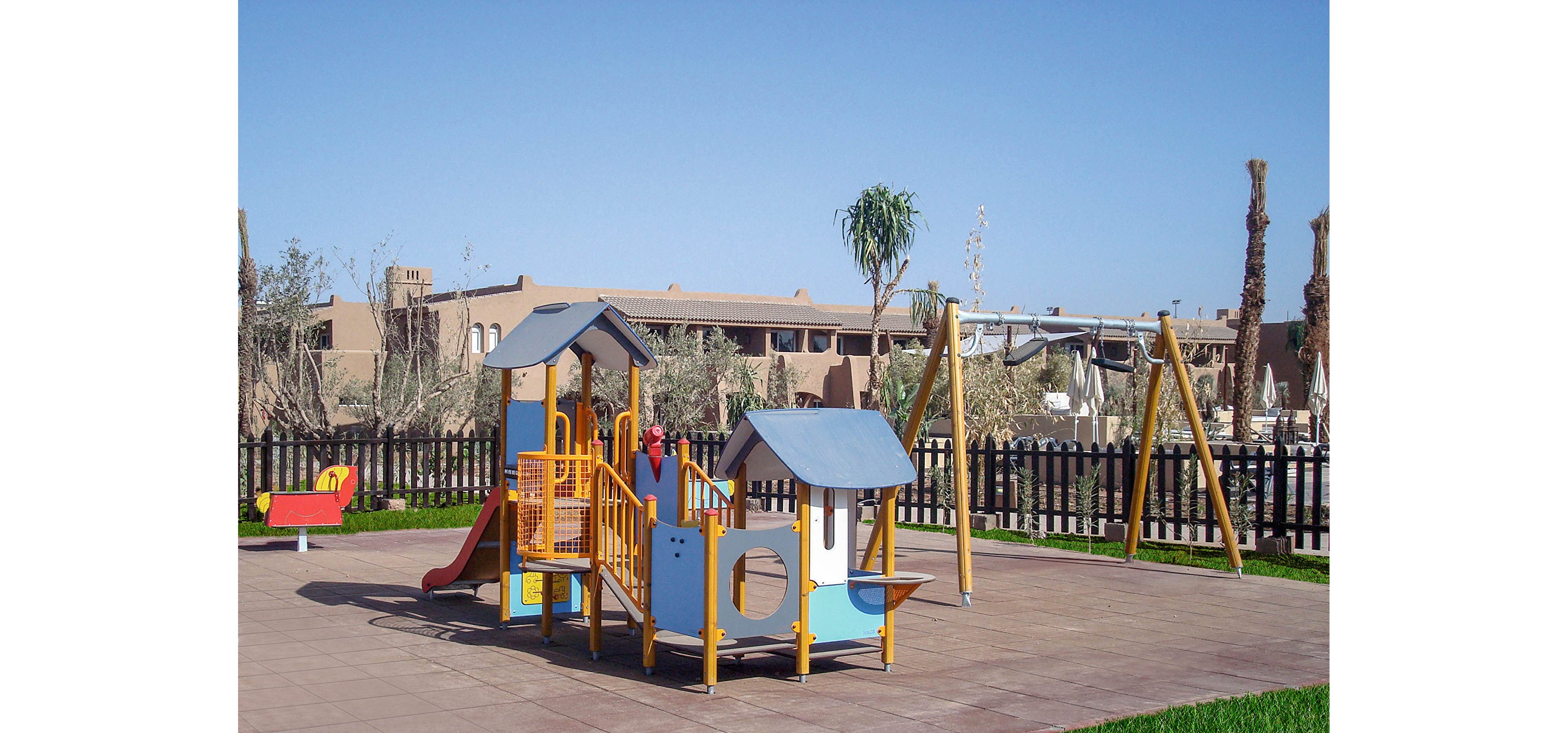 The image shows a colorful playground structure featuring slides, climbing elements, and swings. The setting is an outdoor park with a clear blue sky and surrounding buildings in the background. The area is paved and fenced, with green grass visible at the edge. No people are present, and the mood is bright and inviting.