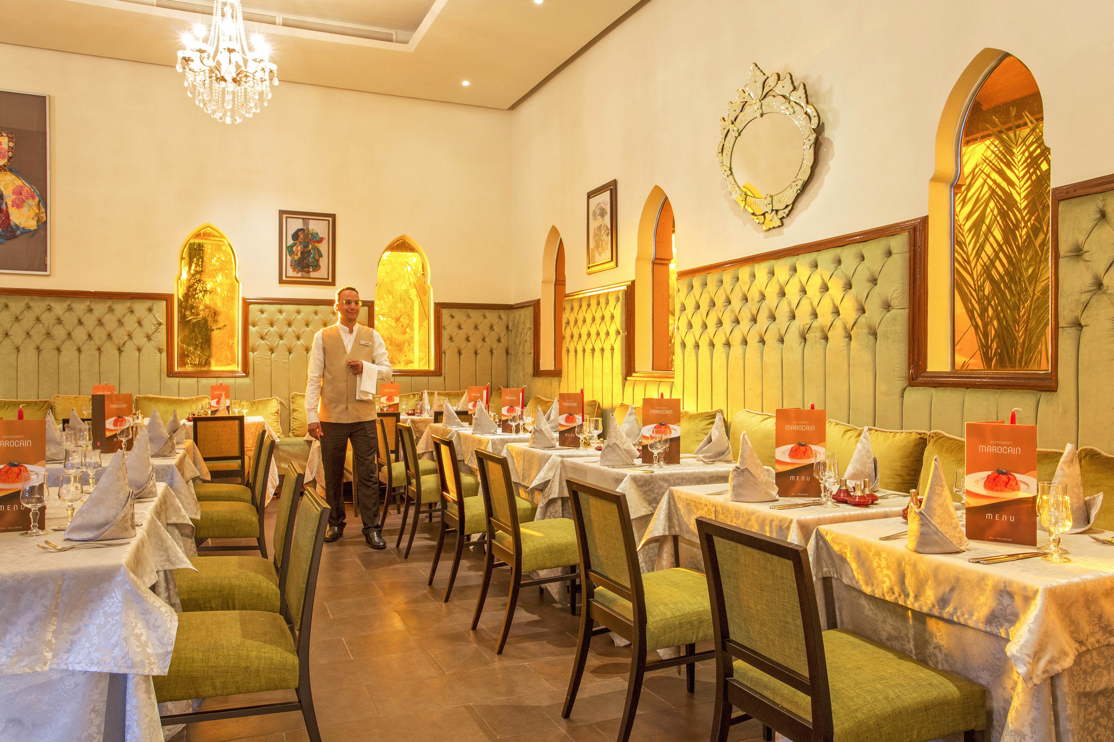 A well-lit restaurant dining area features neatly arranged tables with white tablecloths, folded napkins, and glassware. A male waiter stands in the center, ready to serve guests. The decor includes tufted green wall panels, arched mirrors, and warm lighting, creating a welcoming ambiance. Menu cards are visible on each table, adding a touch of sophistication. The overall mood is inviting and refined.