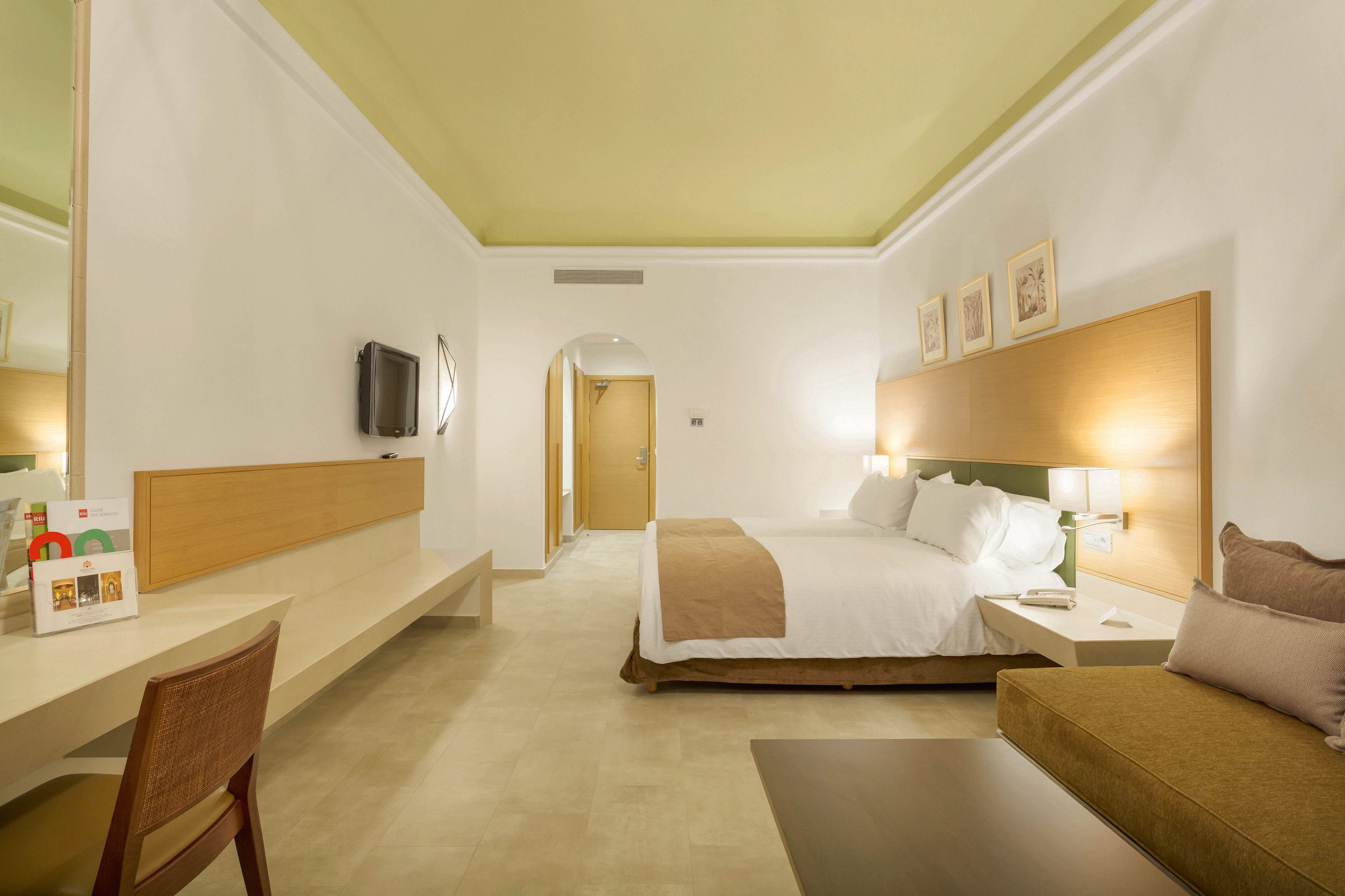 A spacious hotel room features a large bed with white linens and beige accents, a wall-mounted television, and a comfortable seating area. The setting is clean and minimalist, with light-colored walls and flooring, and soft lighting creating a welcoming atmosphere. Artwork is displayed above the bed, and a desk with a chair is positioned near a large mirror. No visible numeric values or identifiable brands are present.