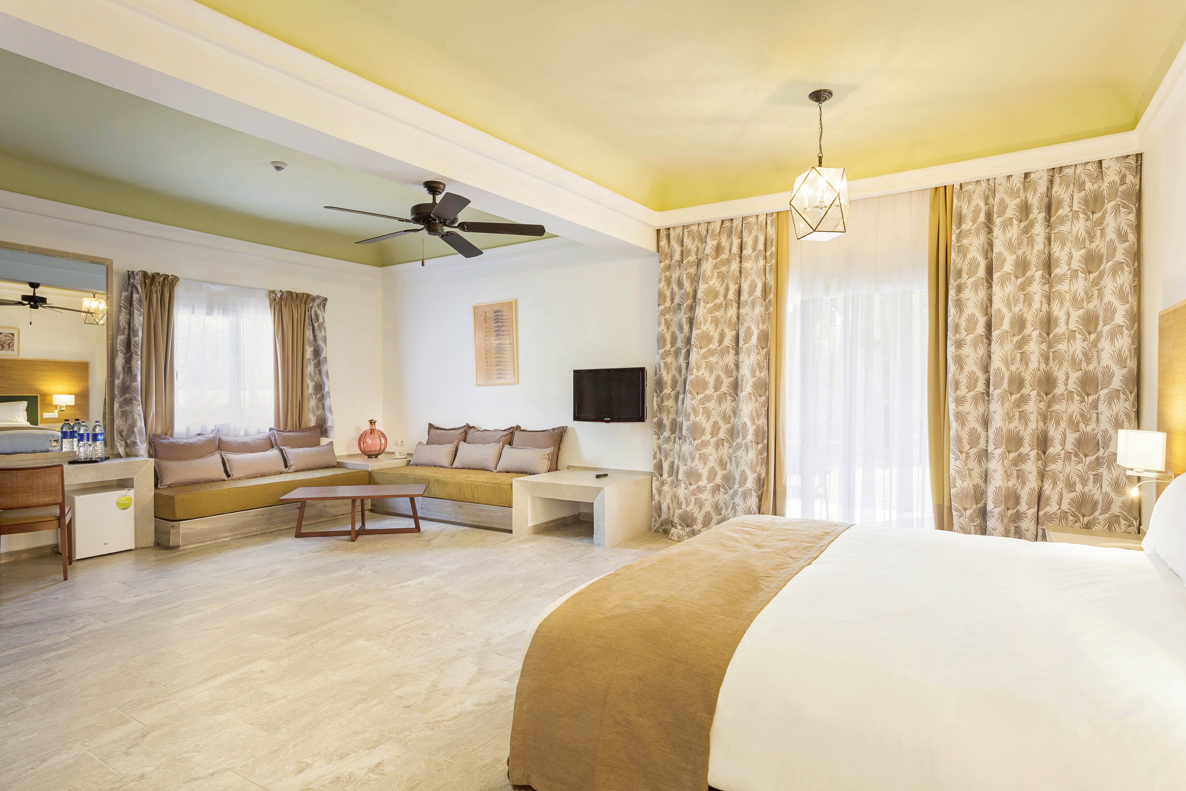 A bright and airy hotel room features a large bed with beige linens, a comfortable corner sofa, and a wall-mounted television. The space is illuminated by natural light through sheer curtains and complemented by neutral tones and patterned drapes. The room includes a ceiling fan, pendant lighting, and a small desk with visible bottled water and glasses.