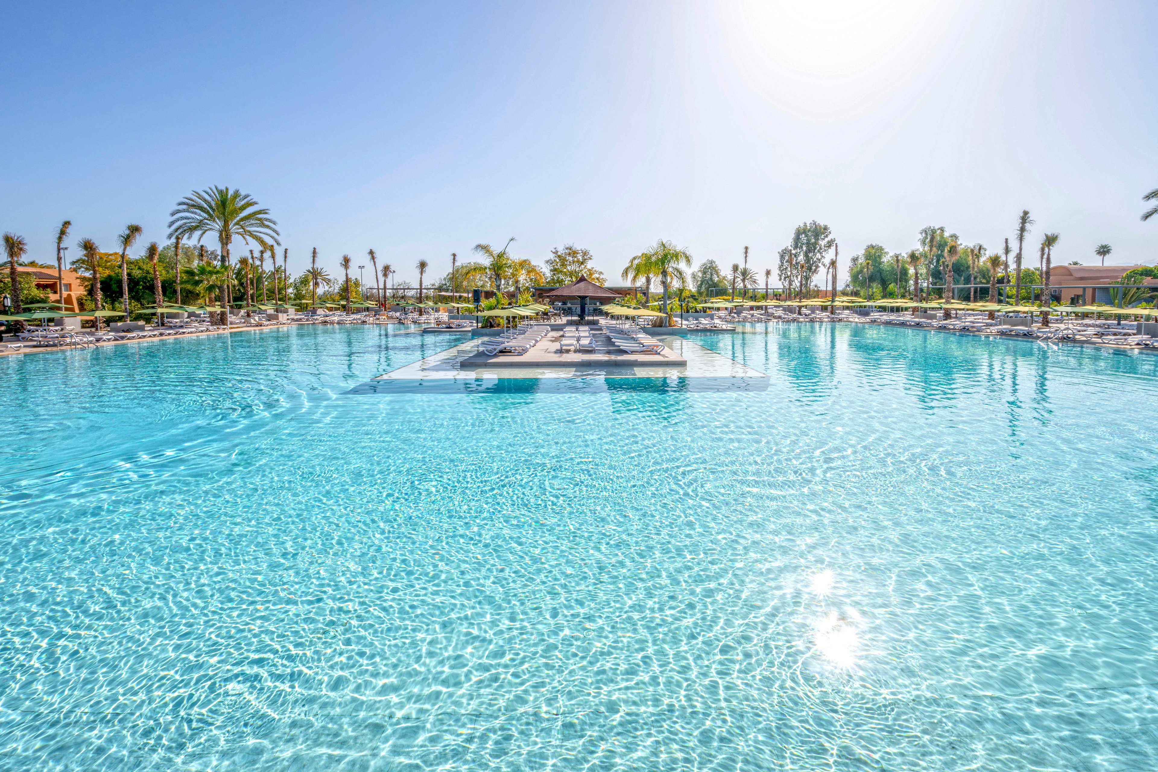 A large outdoor swimming pool is featured under a clear blue sky, surrounded by palm trees and sun loungers. The setting is tranquil and inviting, with sparkling water reflecting sunlight. The central island area in the pool adds visual interest, and the overall mood is relaxed and vacation-oriented. No visible text or numeric values are present.
