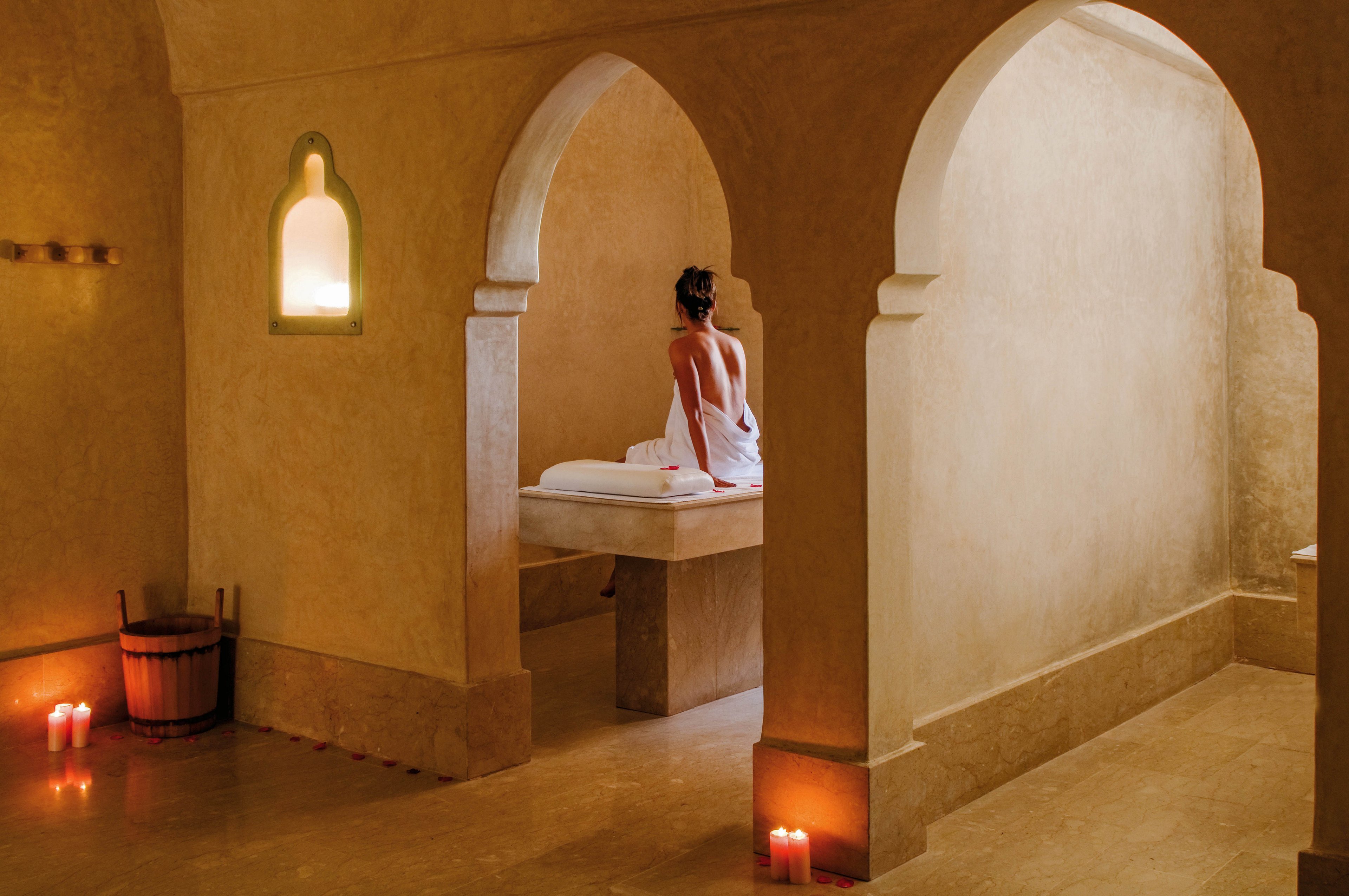 A woman wrapped in a towel sits on a stone massage table in a tranquil spa setting. The room features arched doorways, warm beige walls, and soft candlelight creating a peaceful ambiance. Several red candles and a wooden bucket are visible, enhancing the relaxing atmosphere. The scene evokes calm and wellness in a luxurious environment.
